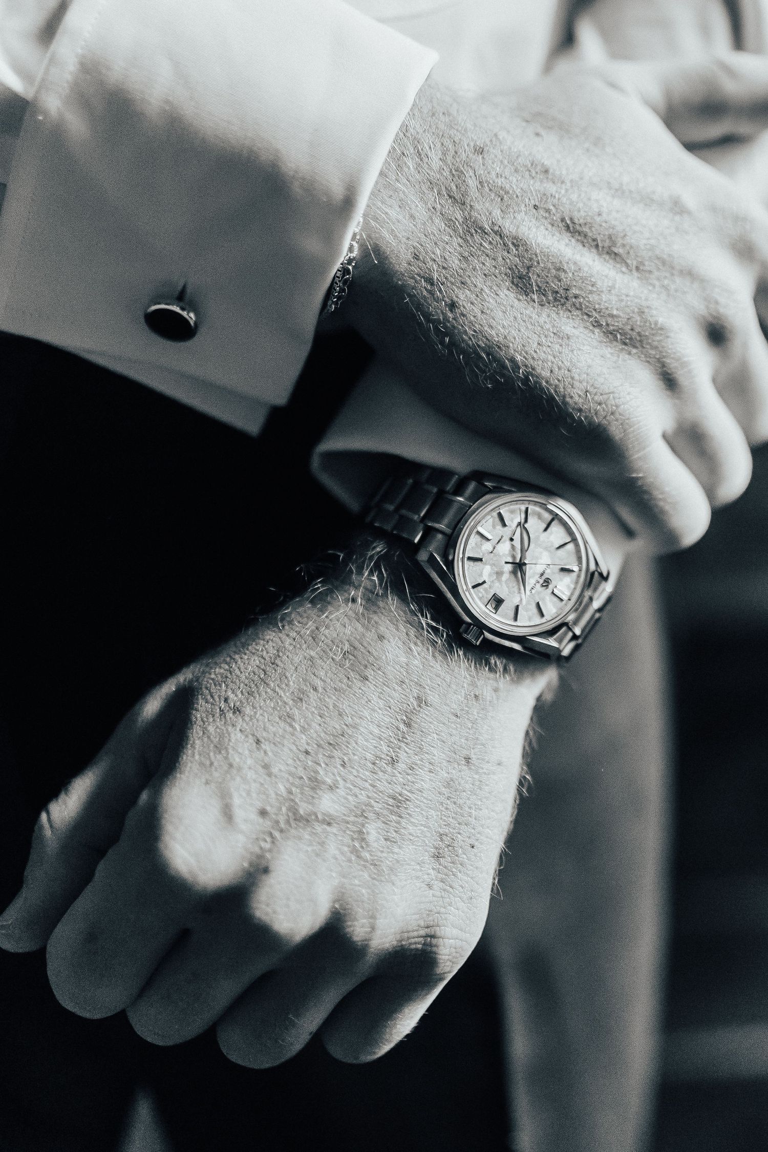 Close-up shot of an elegant wristwatch with leather strap and cufflink visible against a black suit sleeve in monochrome.