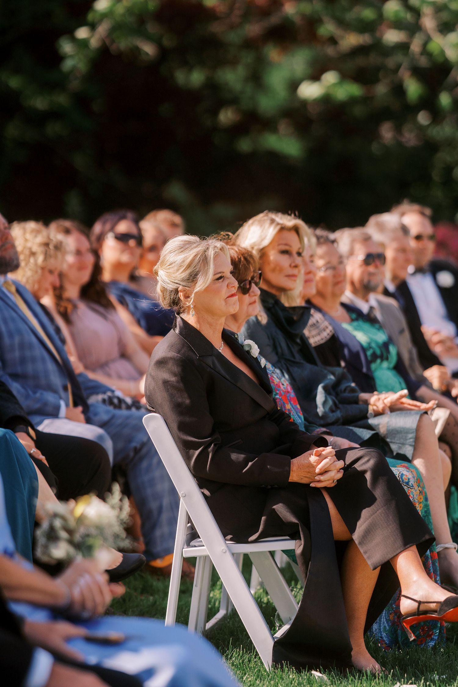 Wedding guests seated in white folding chairs enjoying an outdoor ceremony under tree canopies.