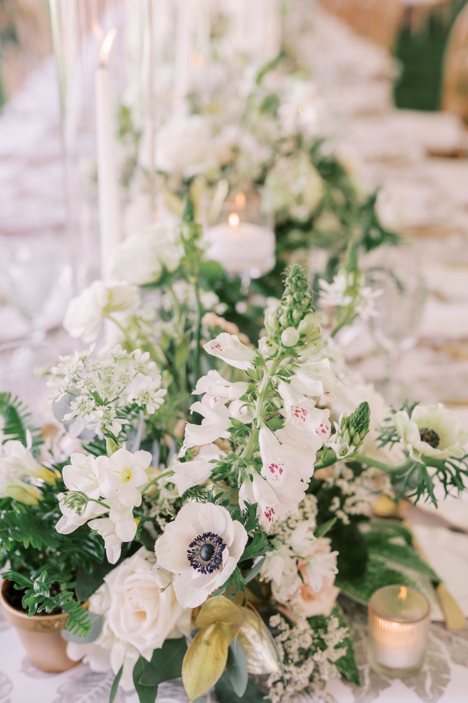 White floral table centerpiece with anemones, roses and greenery alongside glowing votive candles for an elegant wedding reception.