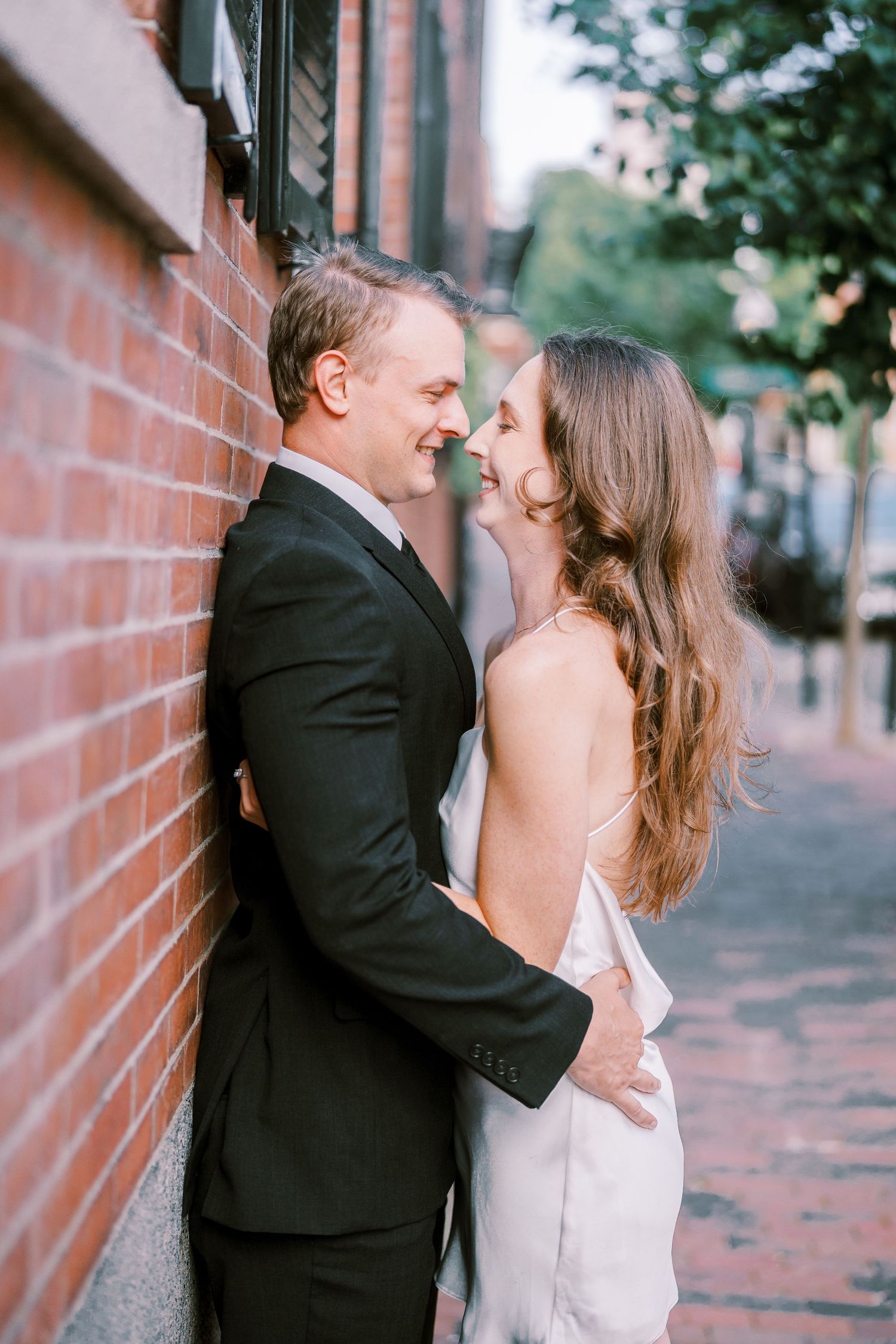 A romantic couple in formal attire embraces against a brick wall on a charming city sidewalk during sunset.