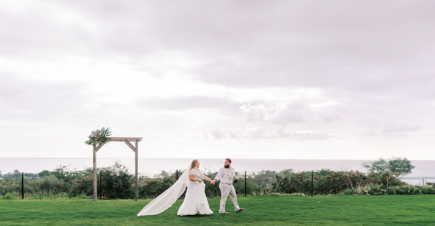 A distant view of newlyweds walking under a wooden arch on a grassy oceanside cliff.