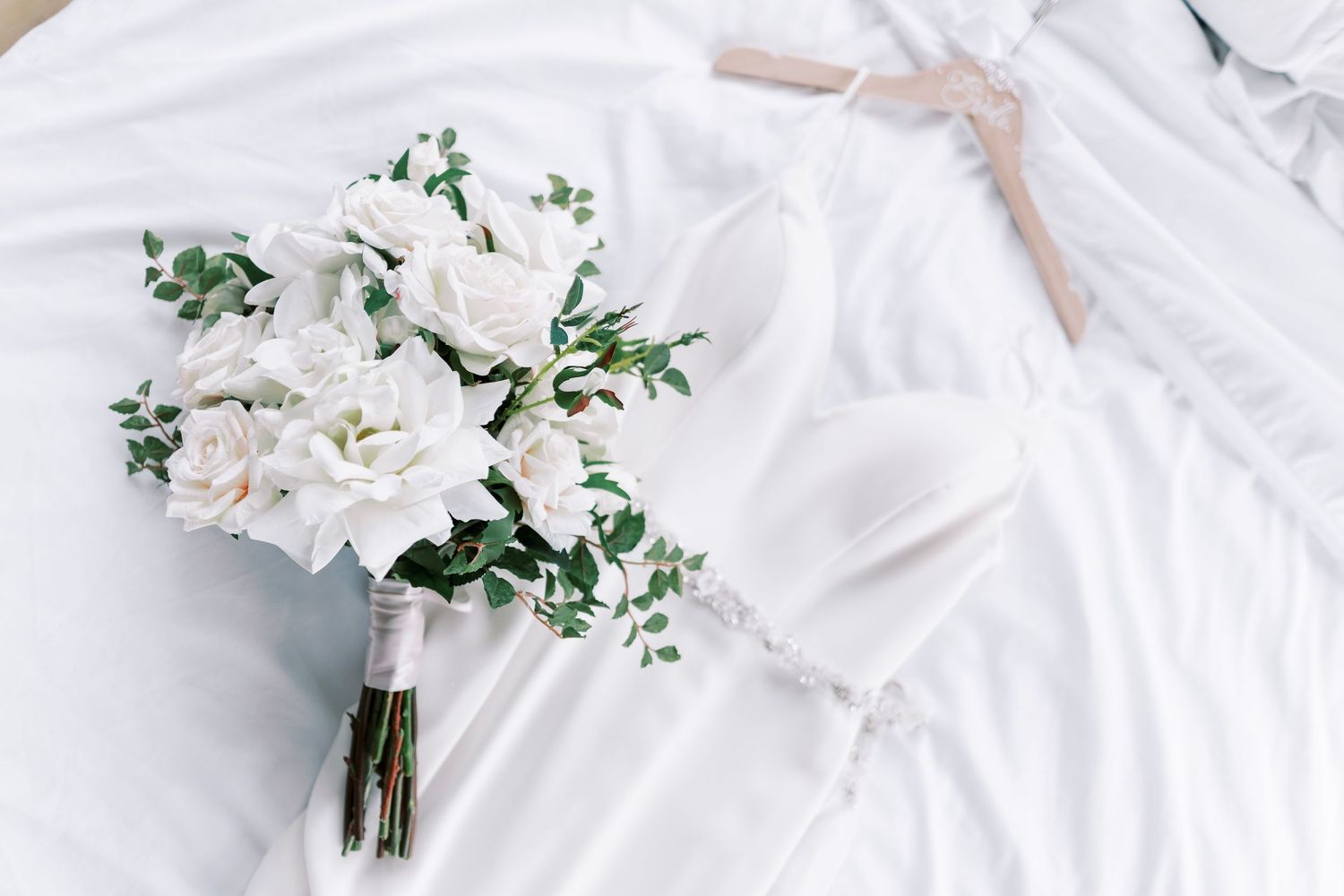A delicate white rose bridal bouquet with eucalyptus greenery rests beside a wedding dress on white fabric.