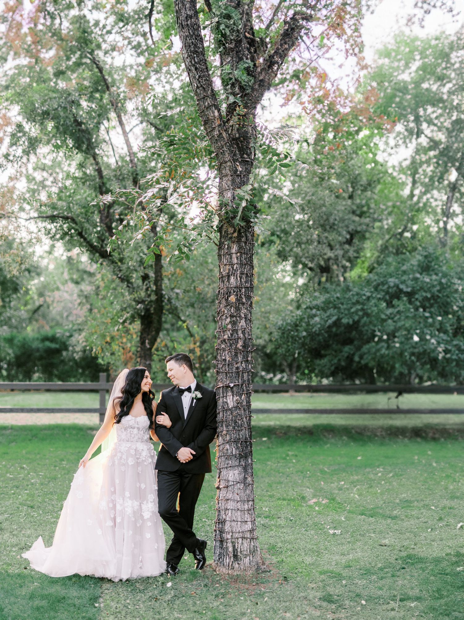 A romantic outdoor wedding photo session showing a couple walking hand in hand under tree branches in a serene park setting.