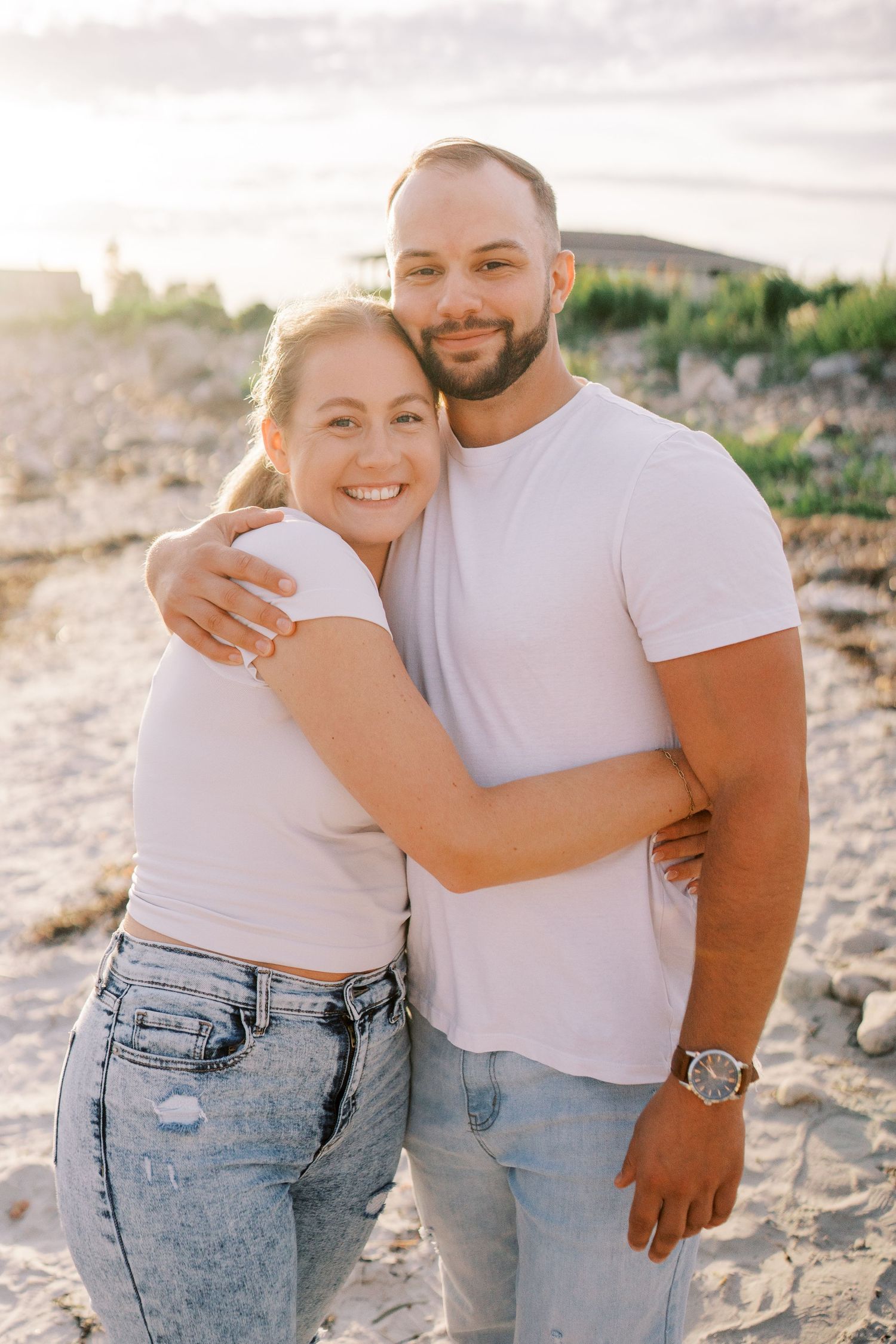 Couple in white shirts and denim jeans pose together on sandy beach at sunset.