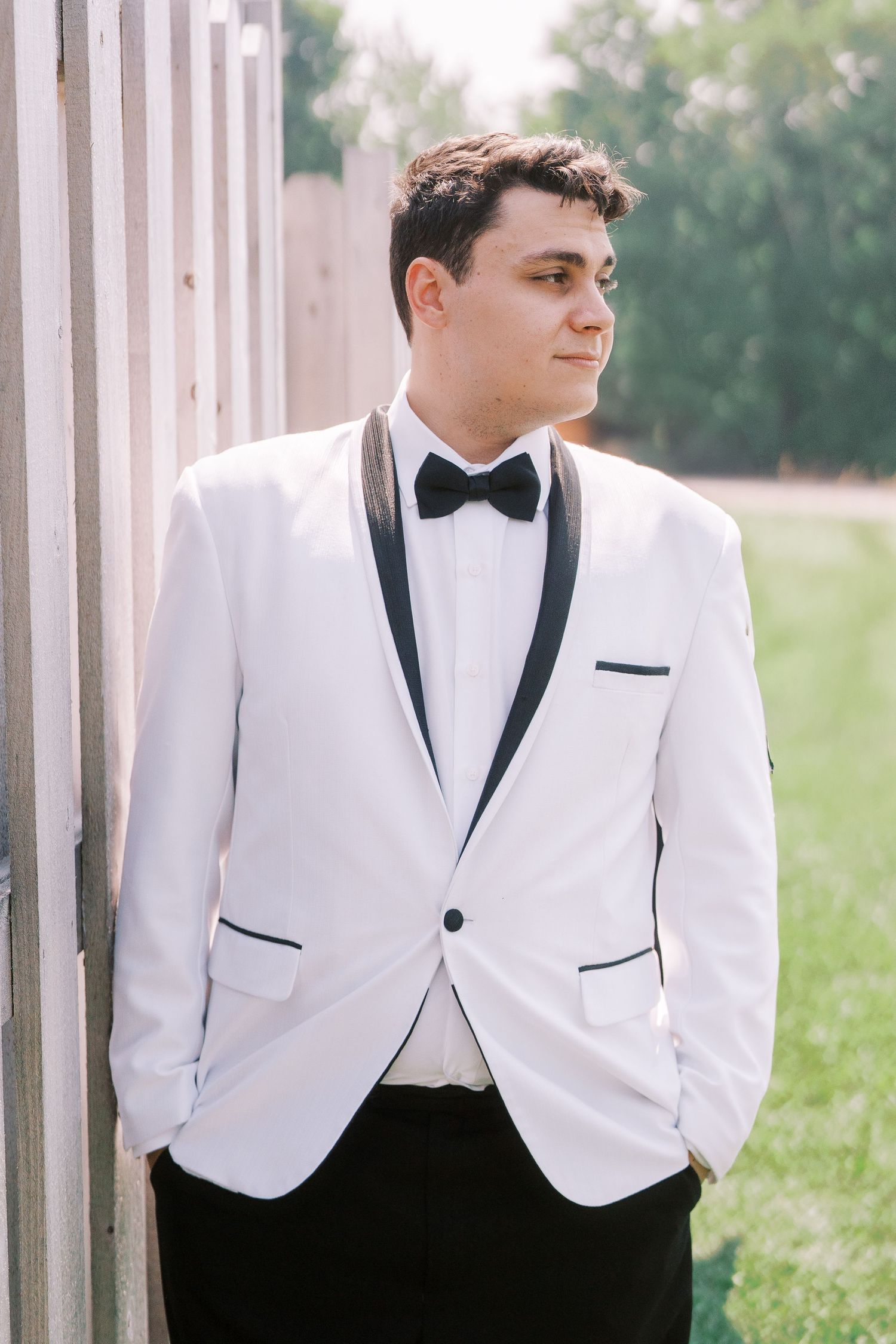 A well-dressed person in a classic white tuxedo jacket with black lapels and bow tie leans against a pillar outdoors.