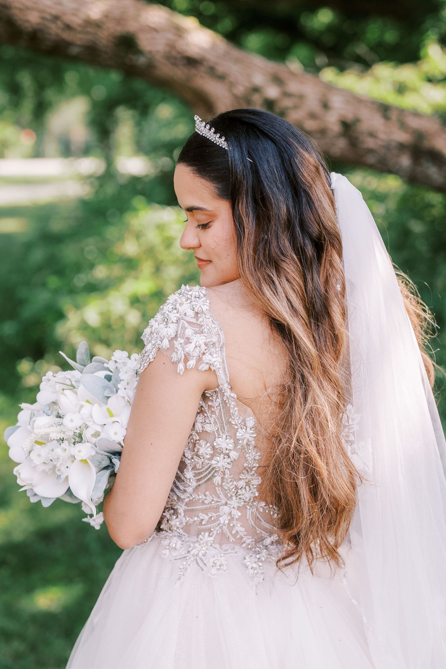 Bride in delicate lace wedding dress with long veil holds white orchid bouquet in outdoor garden setting.