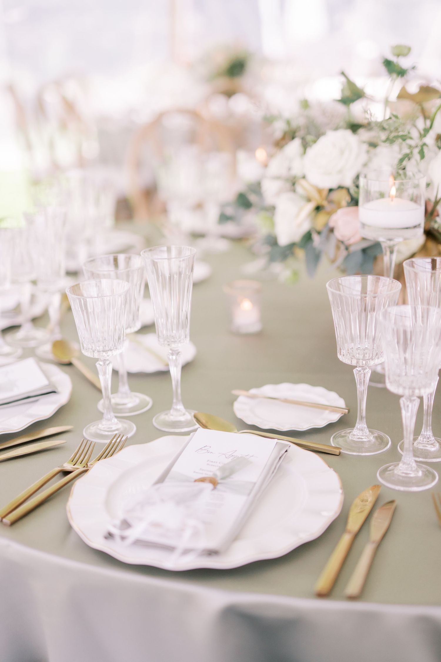 Elegant table setting with crystal champagne flutes, gold cutlery, white plates and soft floral centerpieces on sage green linens.