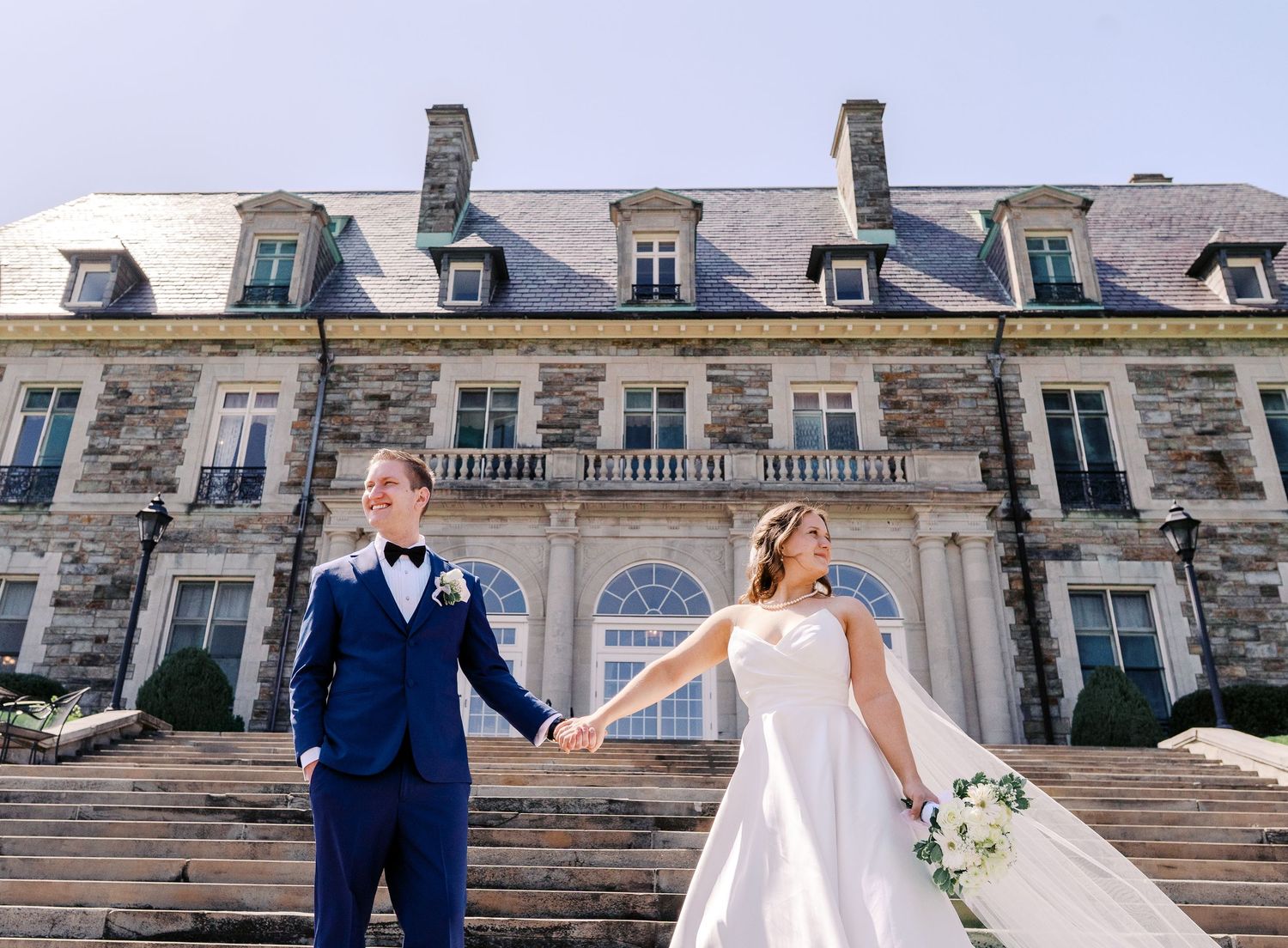 A wedding couple stands hand in hand on stone steps in front of a grand French-style mansion with dormered windows.