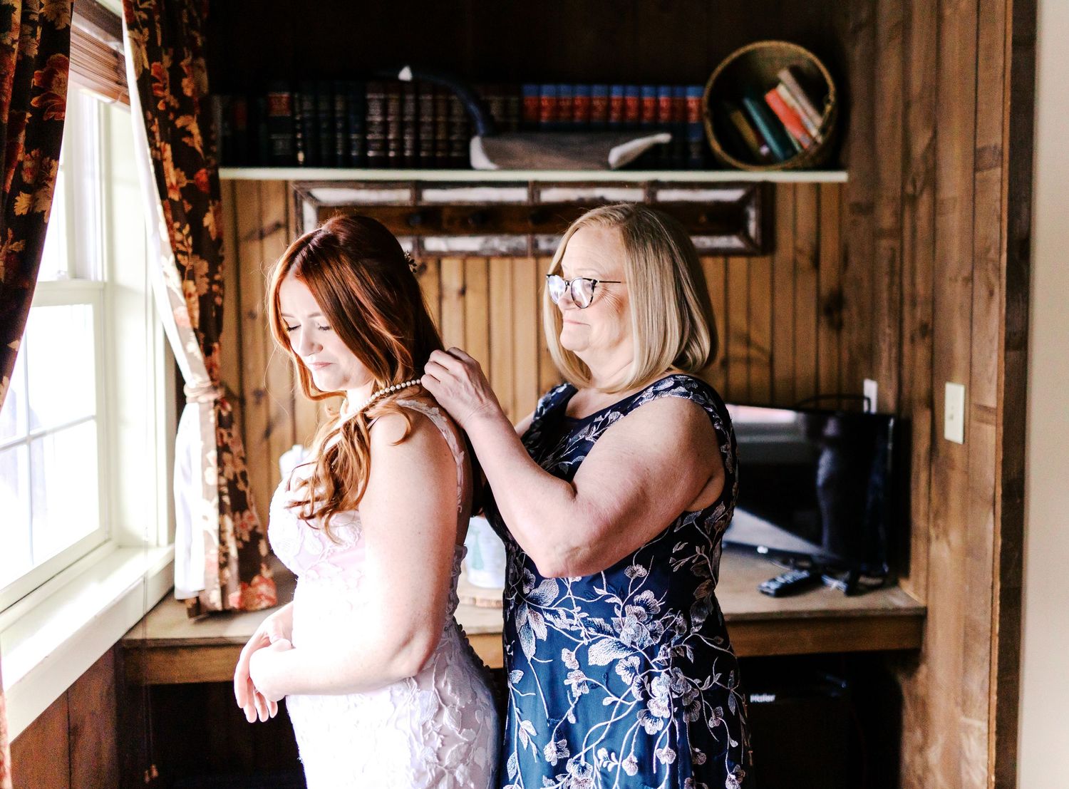 Two women in pink and blue floral dresses stand by a window in a rustic wooden room as one helps the other get ready.