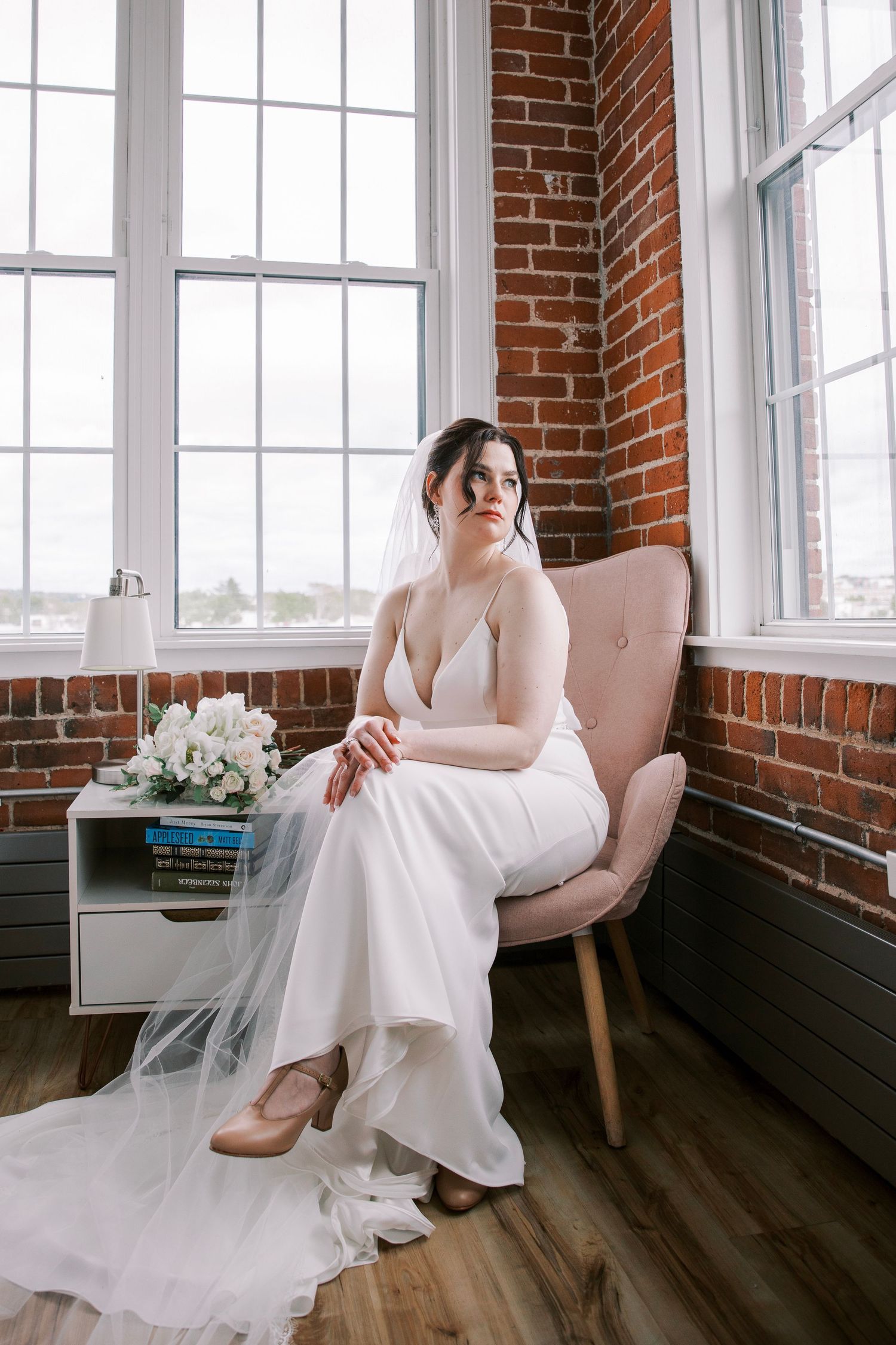 A bride in a white dress sits elegantly on a pink chair by large windows with exposed brick walls.