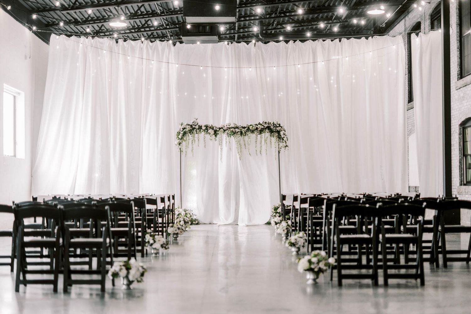 Modern indoor wedding ceremony space with white drape backdrop, black chairs and floral arch decoration in elegant setting.
