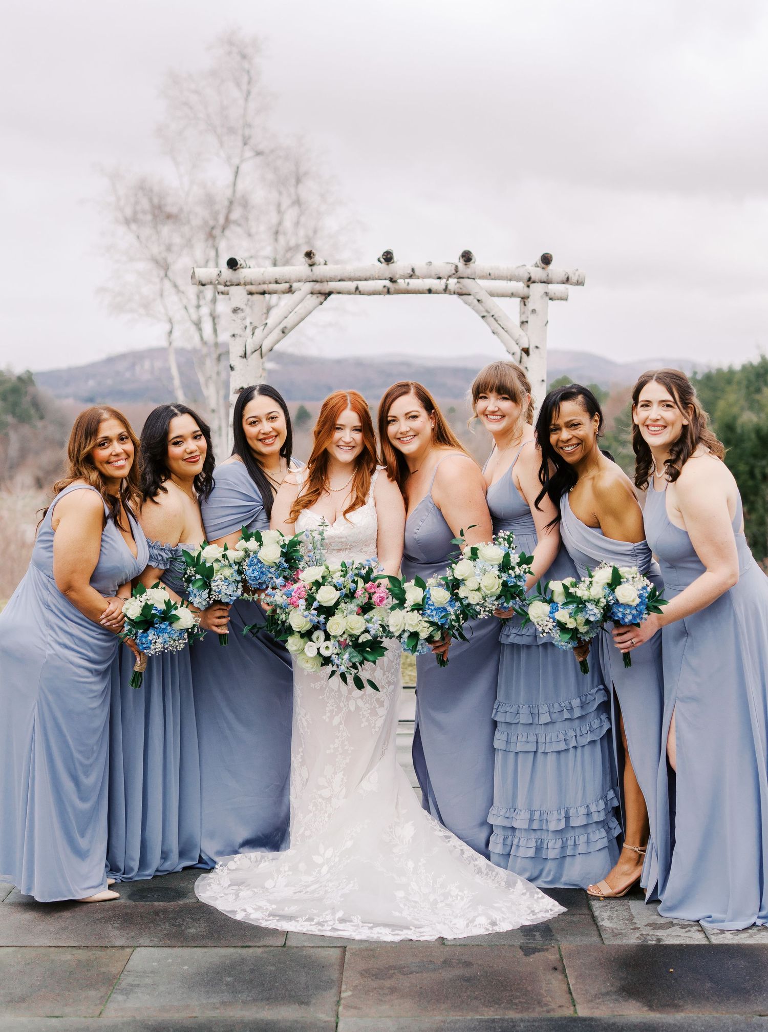 Bridesmaids pose together in dusty blue dresses holding vibrant floral bouquets at an outdoor wedding venue.