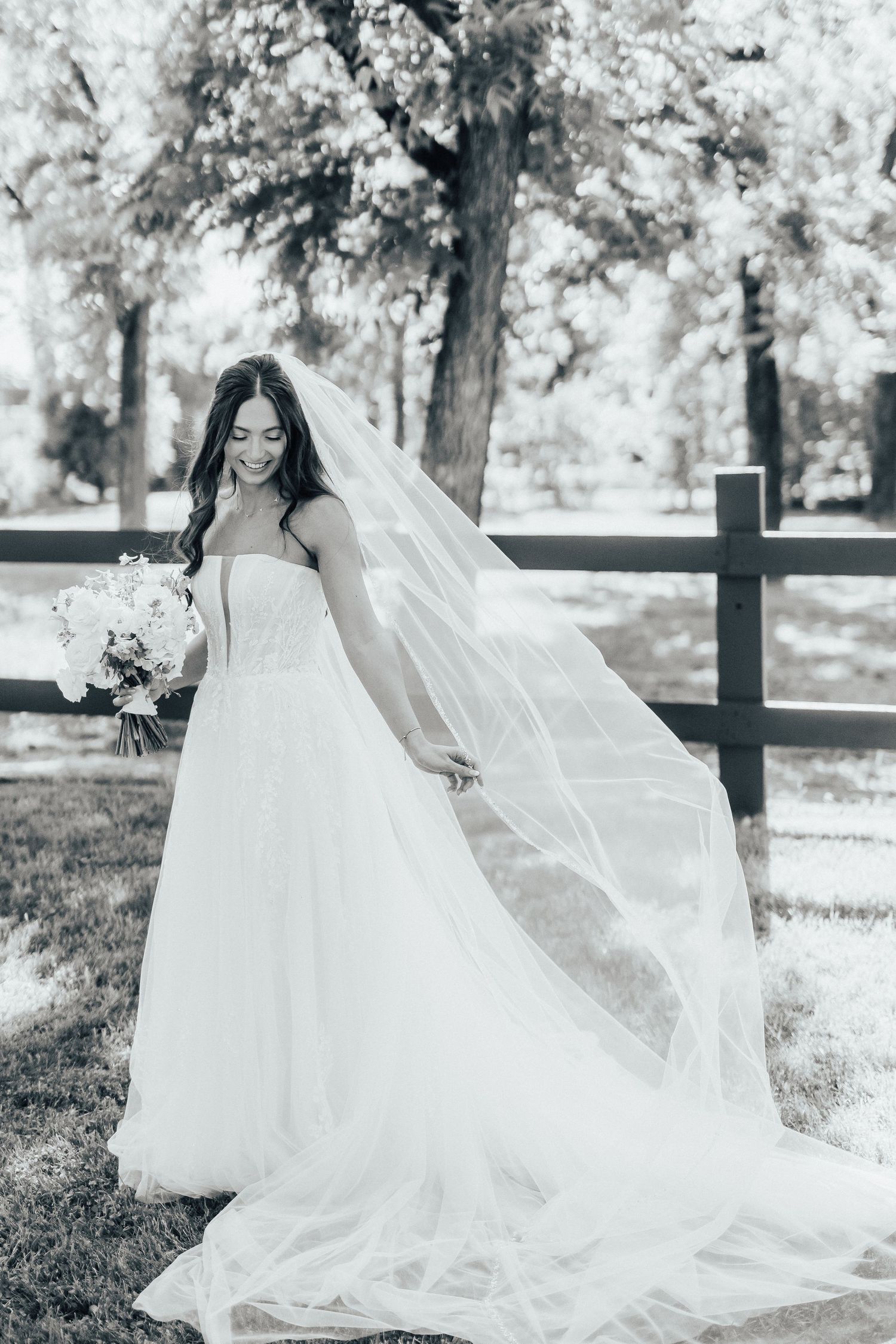 Black and white wedding photo showing a bride in a flowing white dress and long veil posing outdoors by a wooden fence.