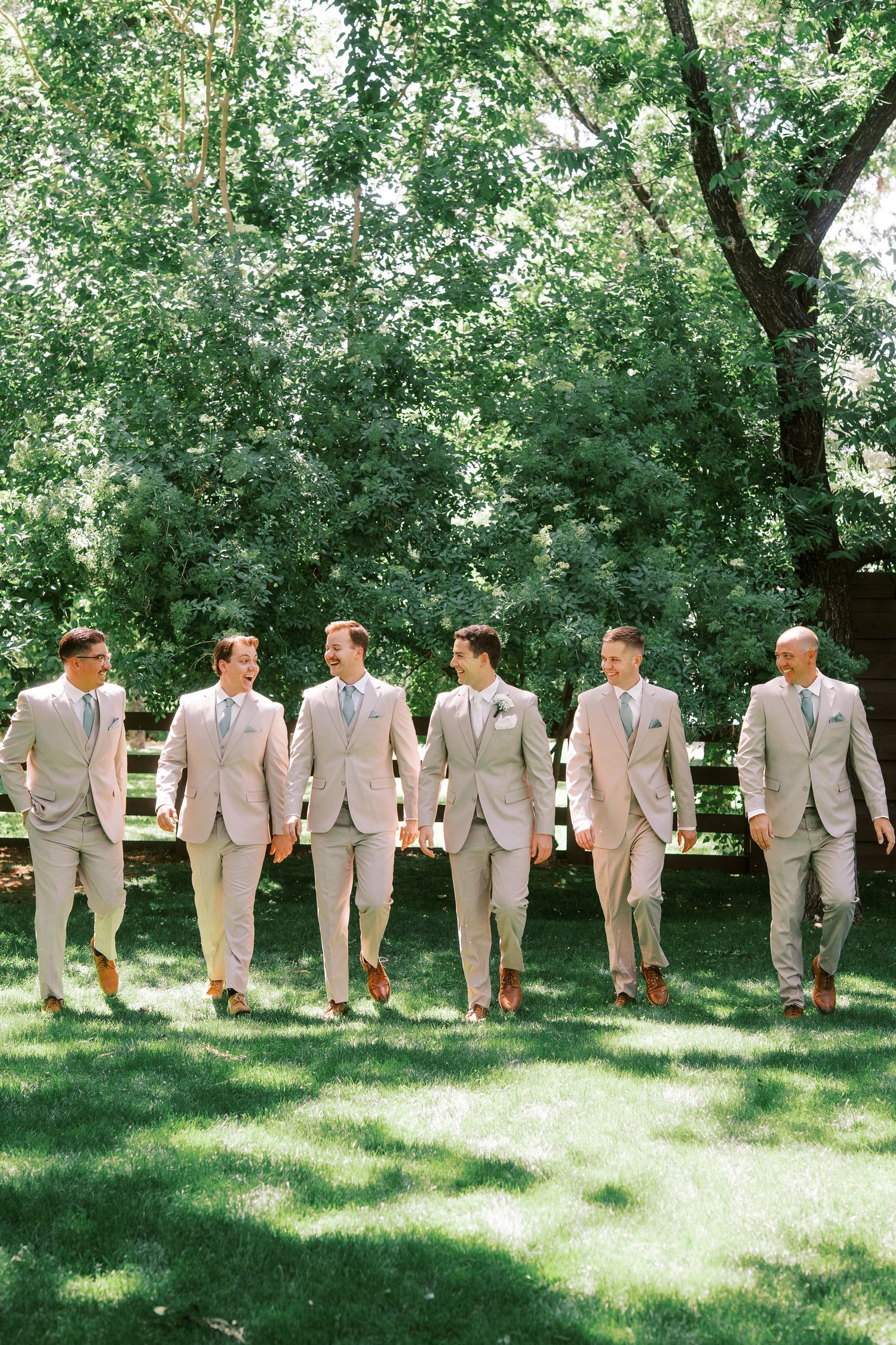 Six groomsmen in light beige suits walking in a line on green grass with trees in the background on a sunny day.
