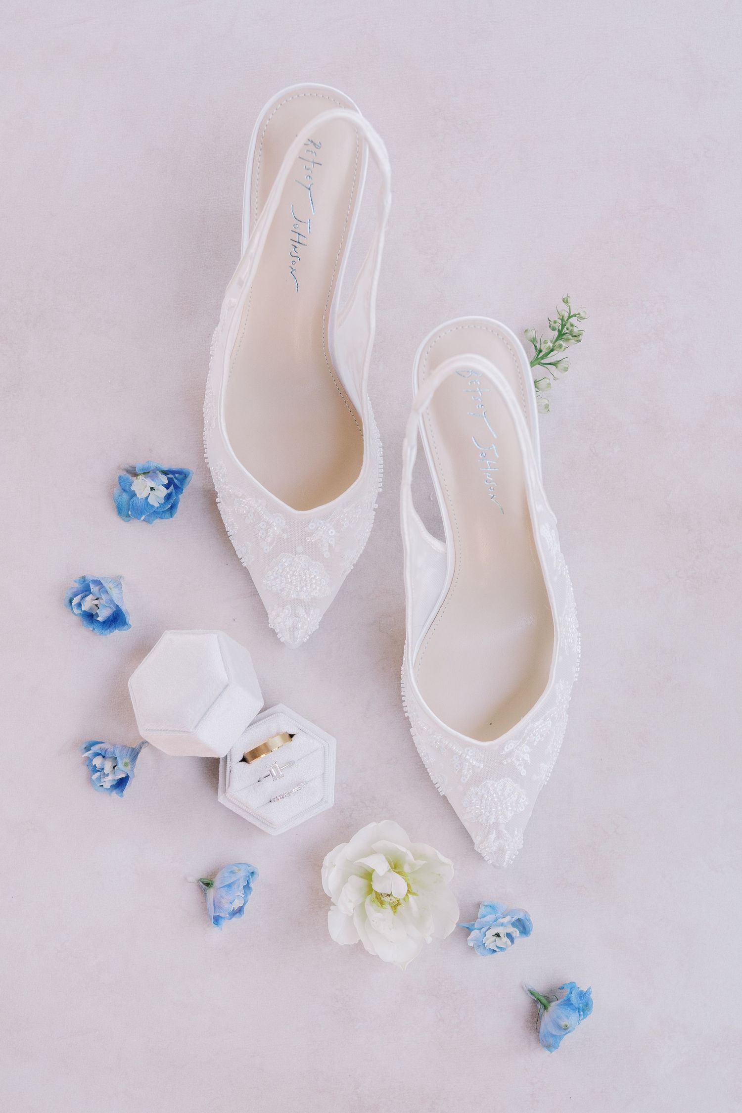 Elegant white lace bridal slingback heels with delicate blue and white floral accents on light pink background.