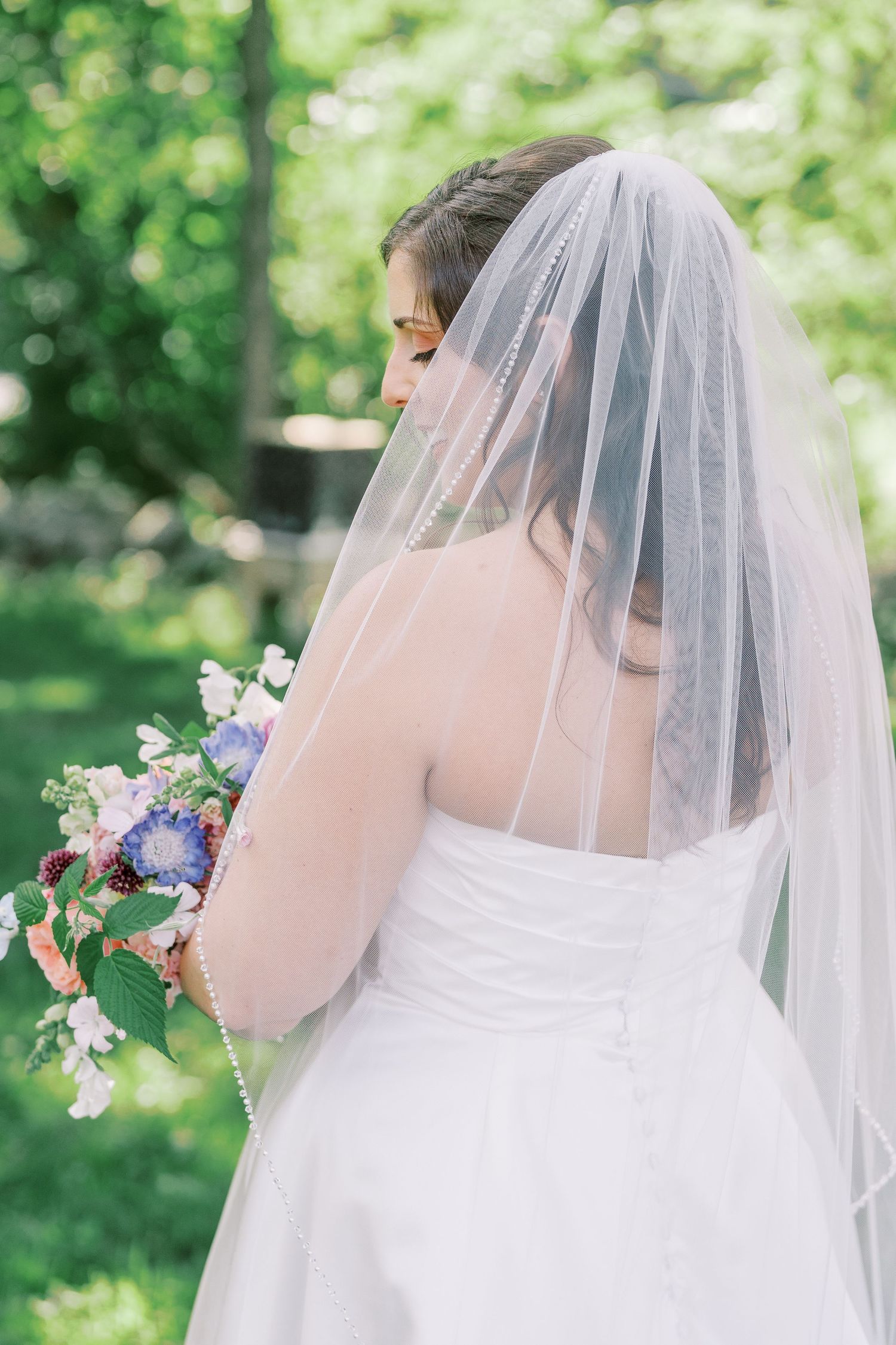 Back view of bride in white dress holding colorful bouquet against green garden backdrop.