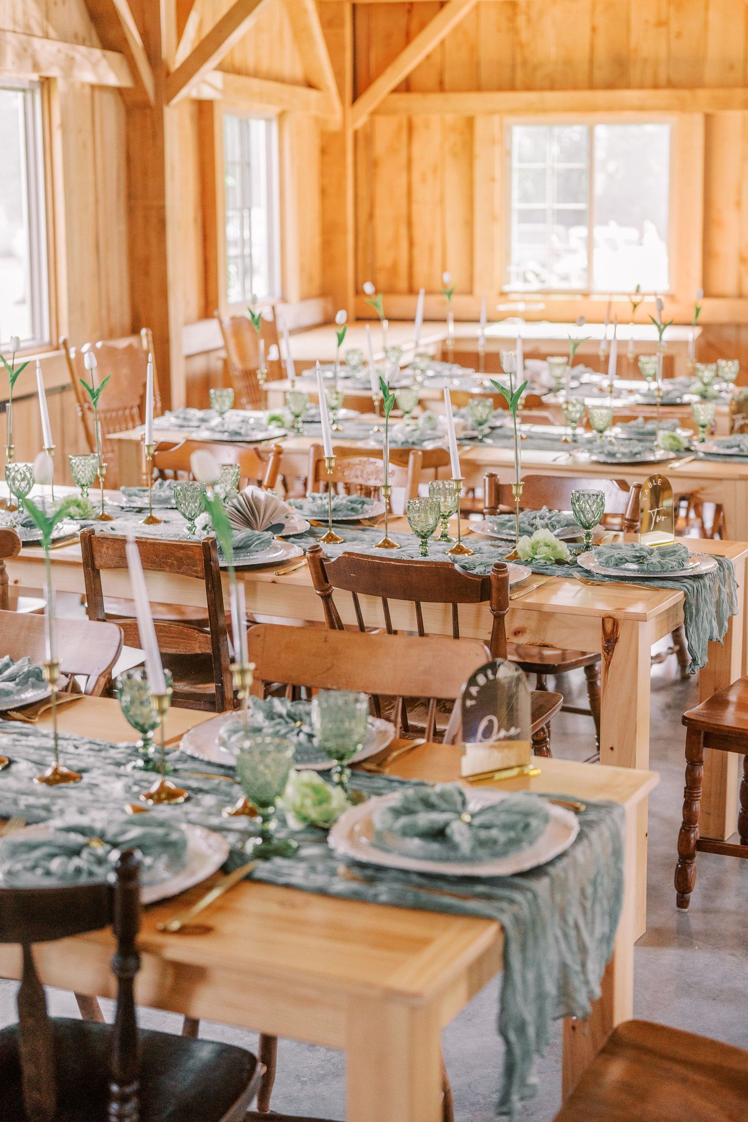 Wedding reception tables set with sage green runners and place settings in a rustic wooden barn venue with natural lighting.