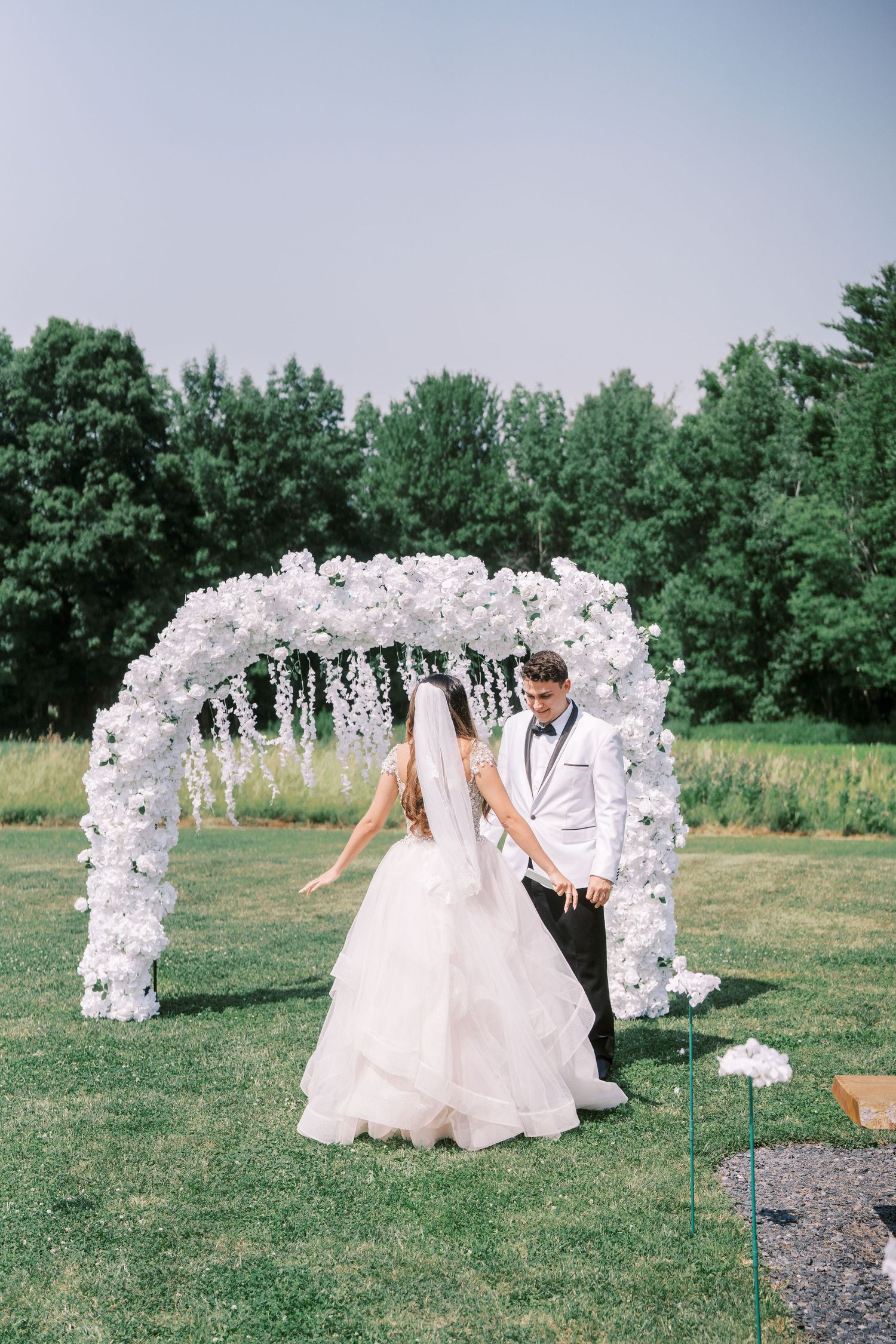 A couple stands beneath a beautiful white floral arch during an outdoor wedding ceremony with green trees in background.