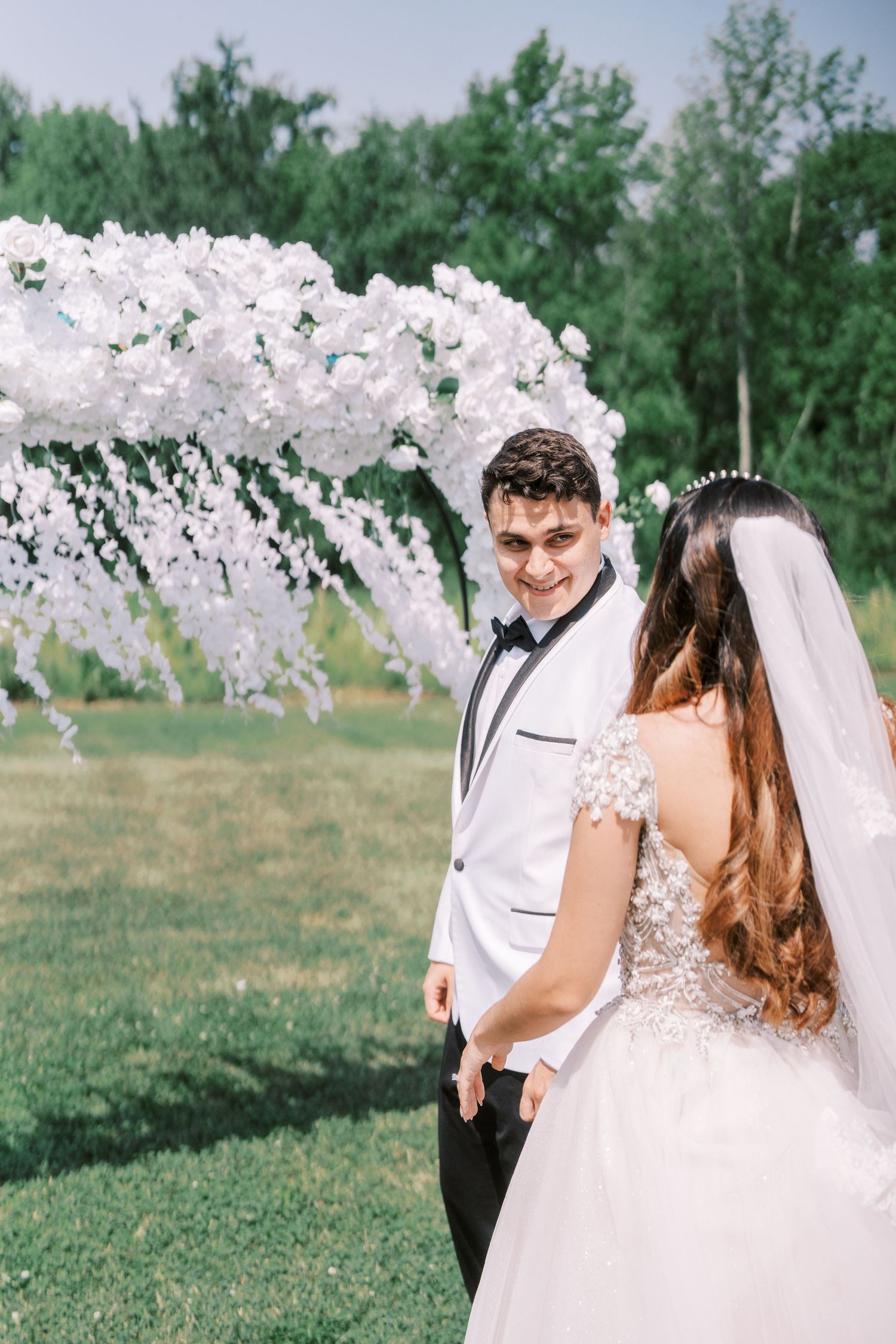 A wedding couple stands together under a beautiful blooming white cherry blossom arch in an outdoor garden setting.