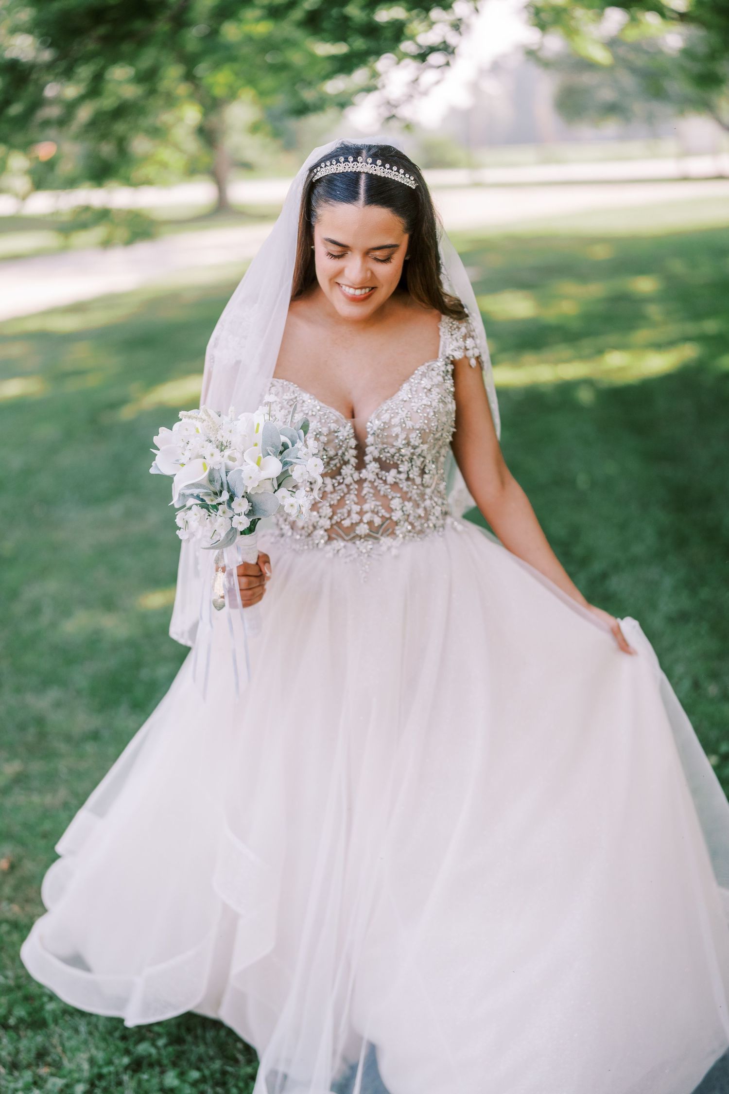 A bride in a white ballgown wedding dress with lace bodice holds her flowing tulle skirt outdoors on a sunny day.