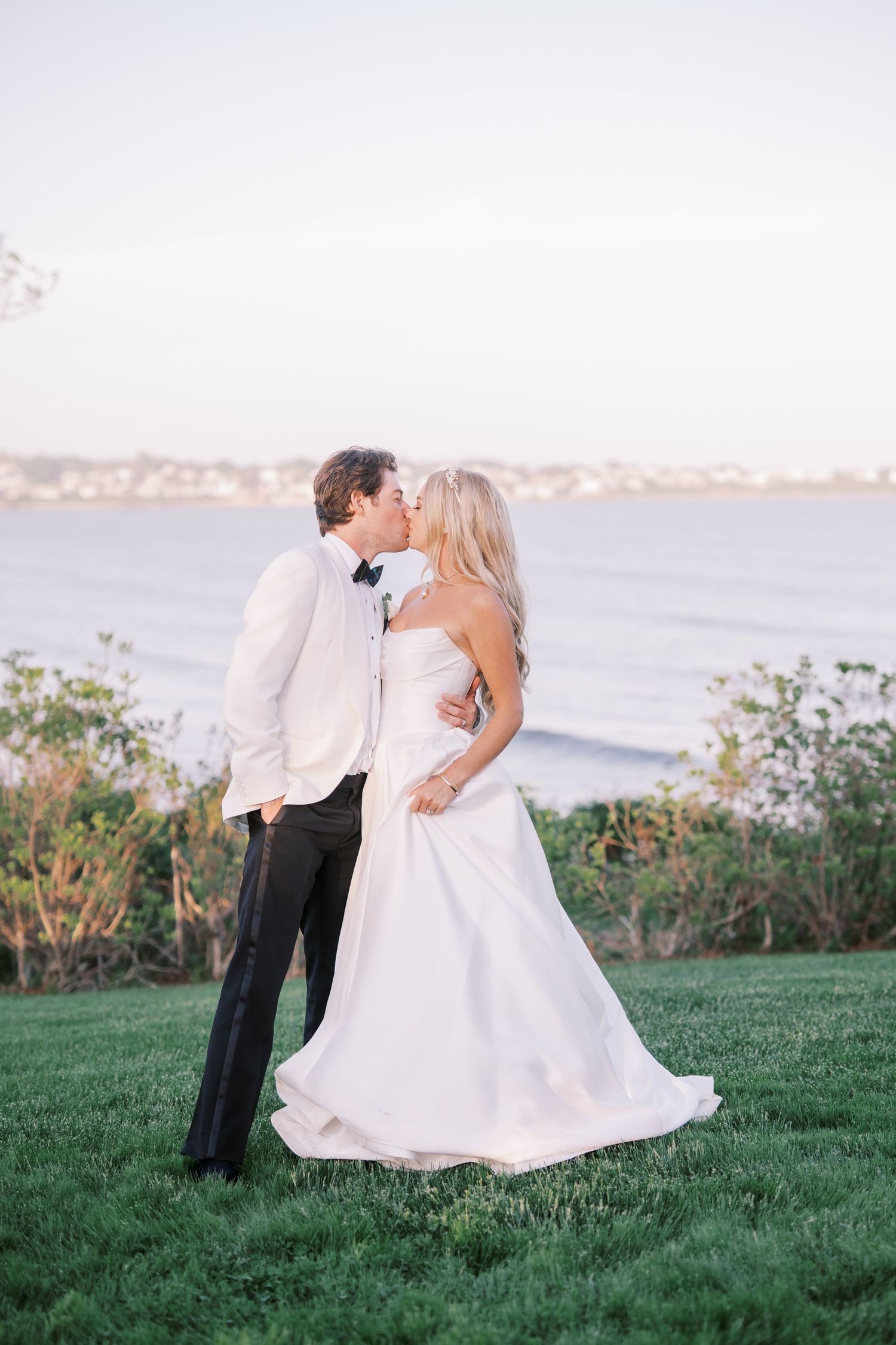 A romantic wedding couple kiss on a grassy oceanside cliff during sunset.