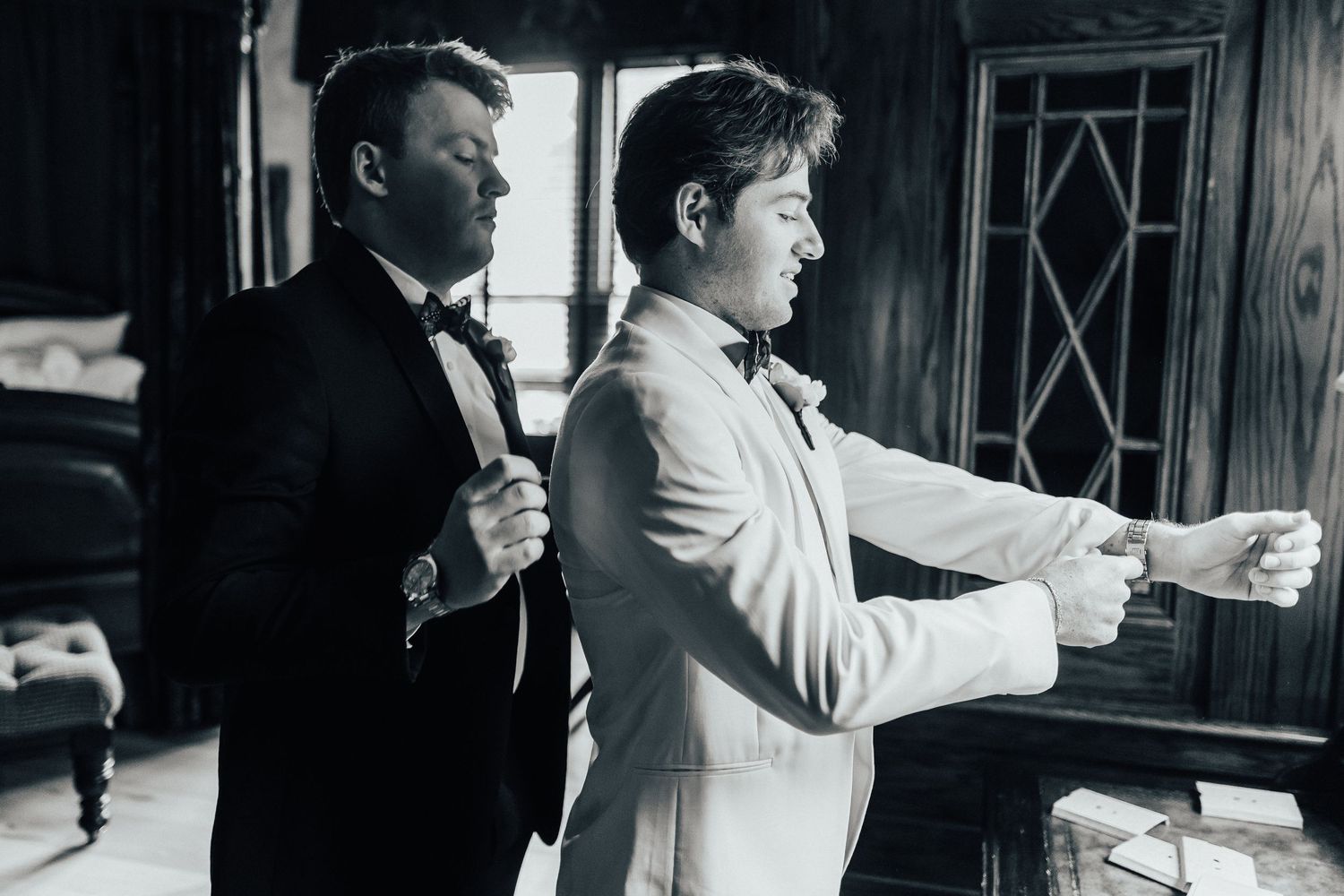 Black and white photo shows two men in formal attire getting ready in a room with ambient lighting through a window.