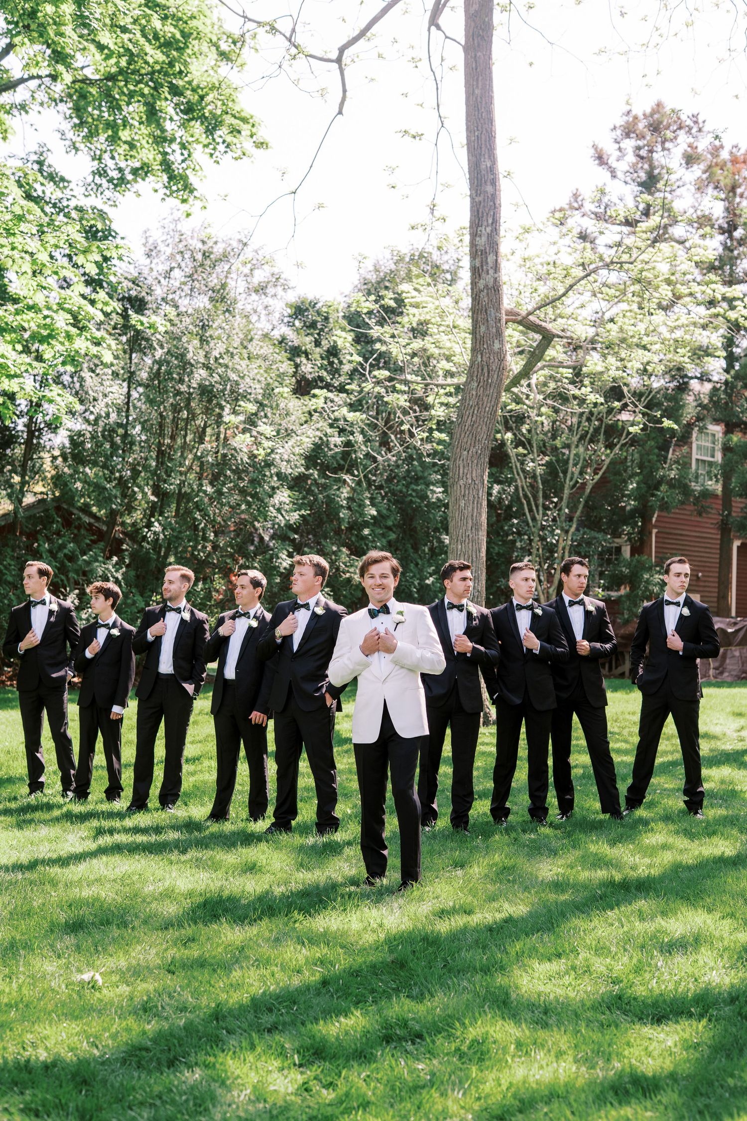 A group in formal attire poses outdoors on a lawn with trees in the background on a sunny day.