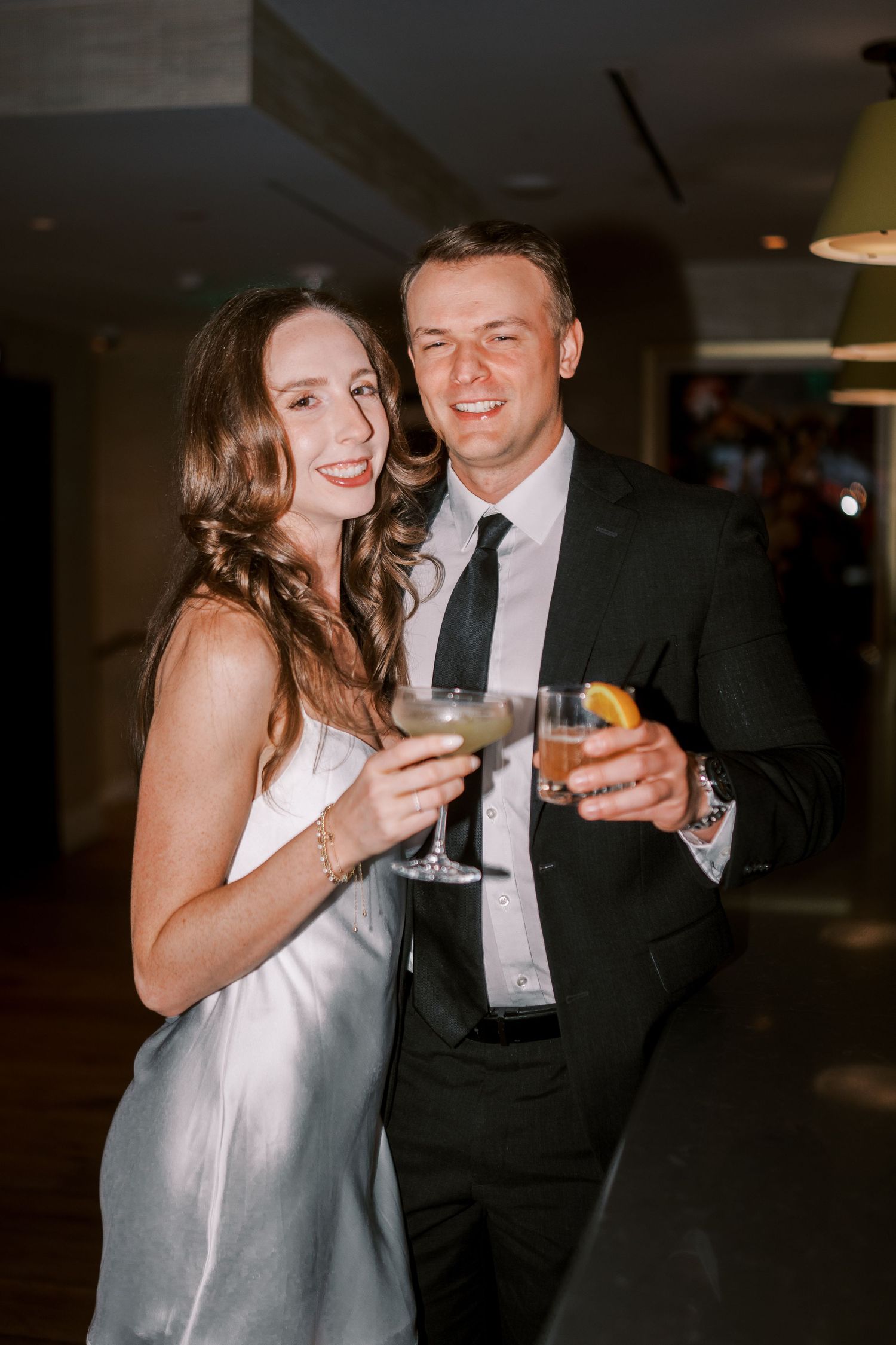 A couple celebrates at an evening event, holding cocktail glasses and dressed in formal attire.