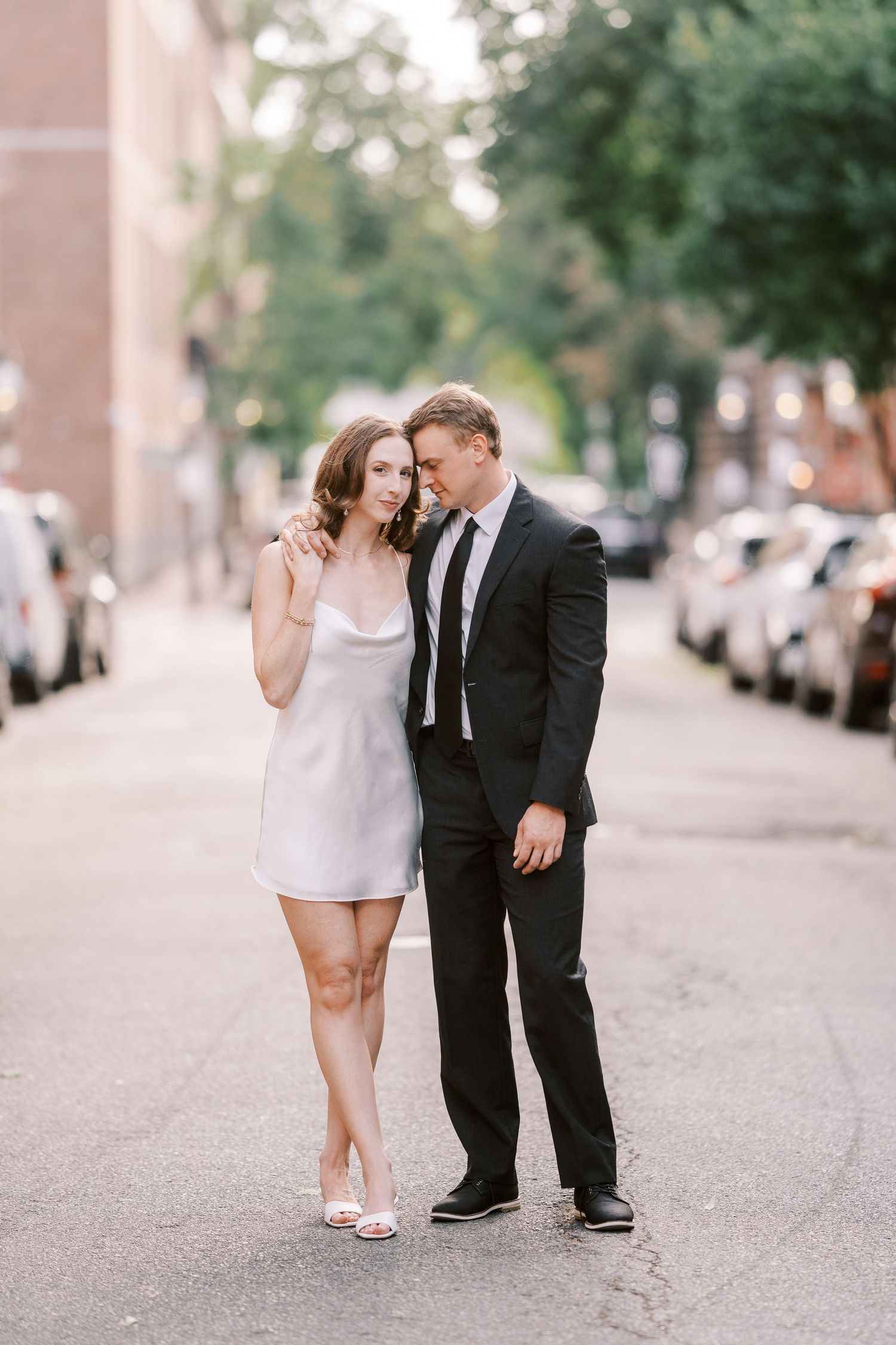 A couple shares an intimate moment on a tree-lined street, with her in a white dress and him in a dark suit.