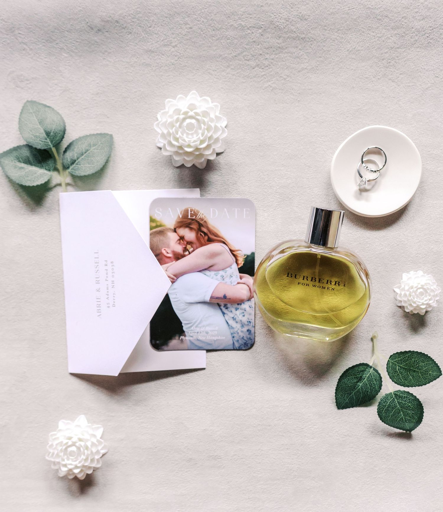 Wedding details flatlay with perfume bottle, rings, and floral decorations on light background.