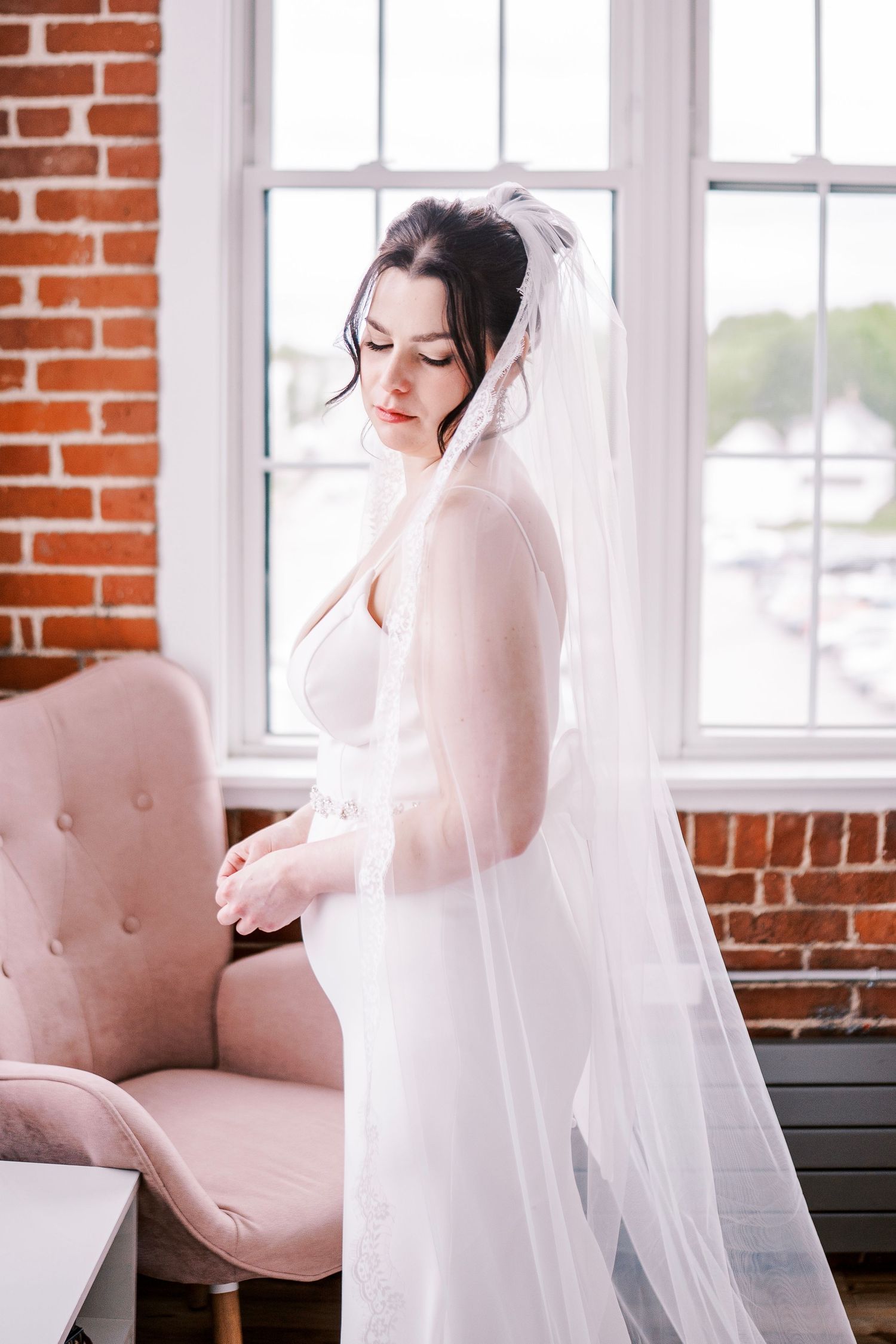 A bride in a sheer white veil sits by a window with exposed brick walls and pink upholstered furniture.