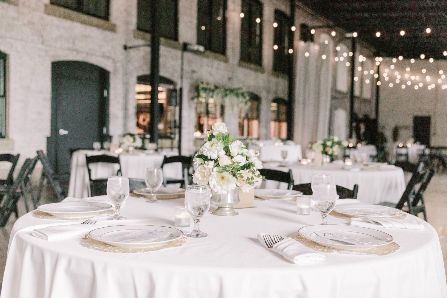Modern industrial wedding venue with round white tables, string lights, and simple floral centerpieces.