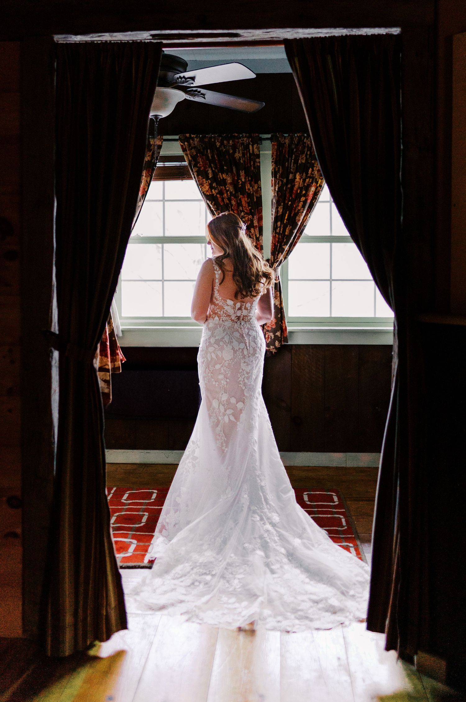 A wedding dress with intricate lace details and a long train photographed dramatically between dark curtains near a window.