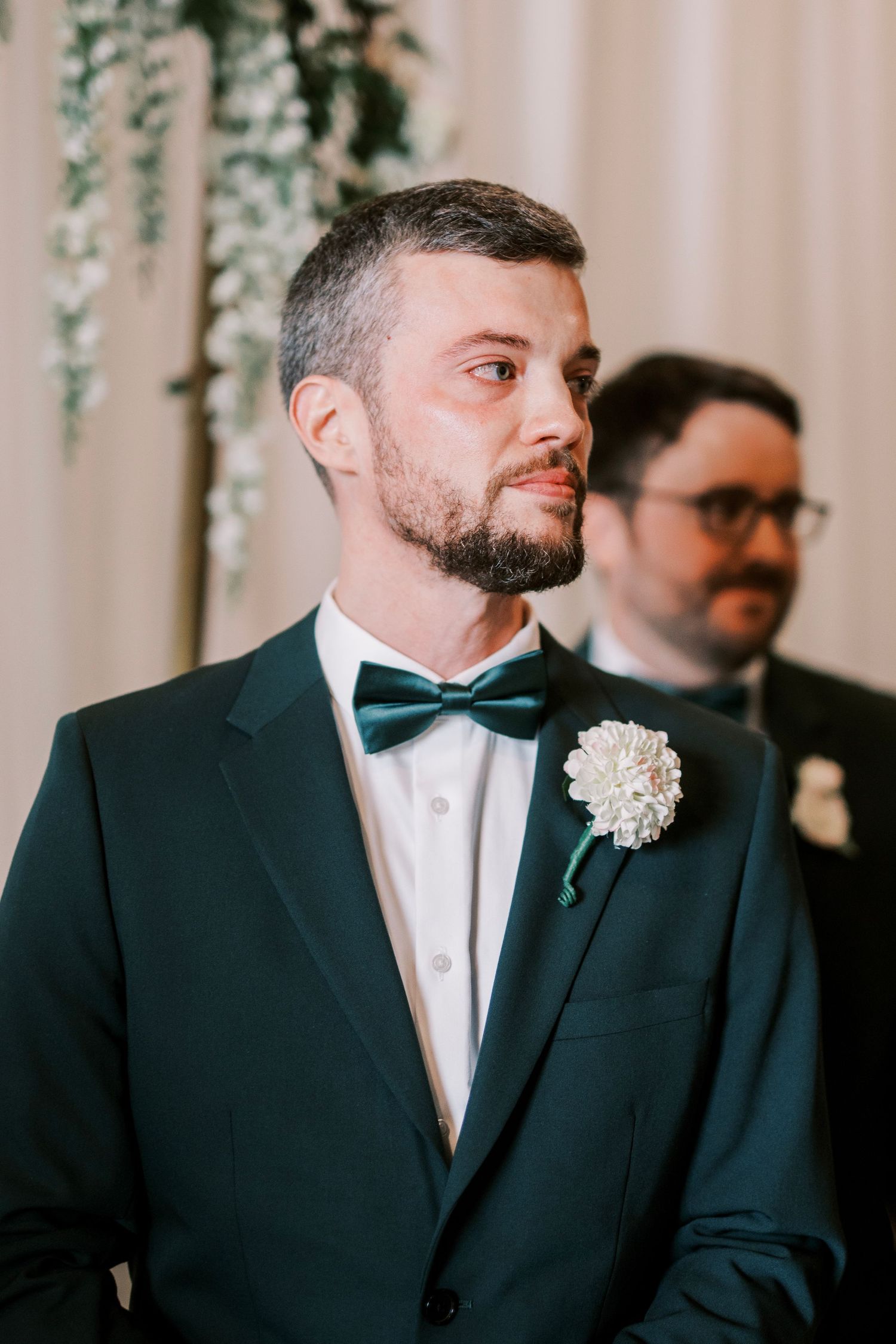 A groom in a classic black tuxedo and bowtie with a white boutonniere looks to the side.