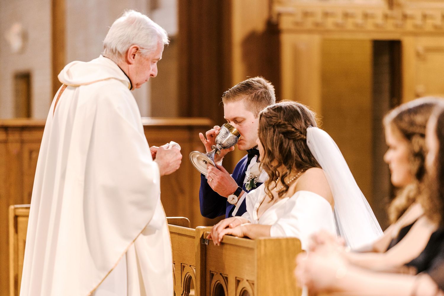 A couple participates in their wedding ceremony as they receive communion from a clergy member in white robes at the altar.