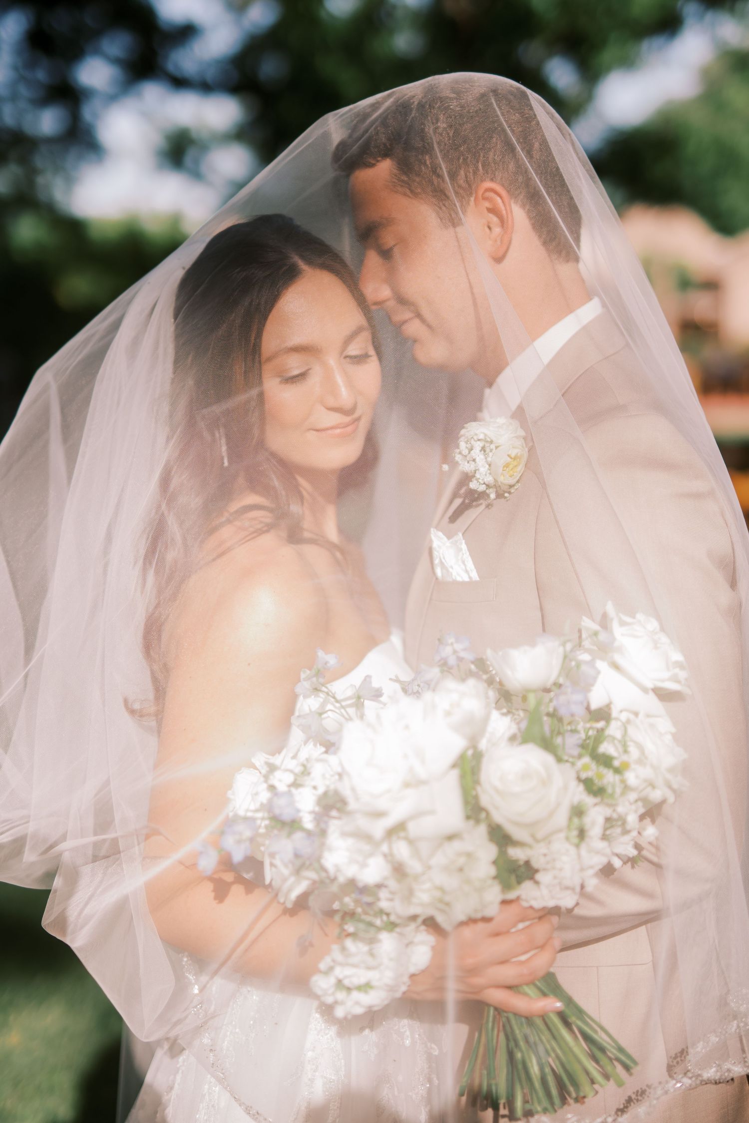 Romantic wedding couple poses beneath a flowing white veil in a garden setting, holding a white floral bouquet.