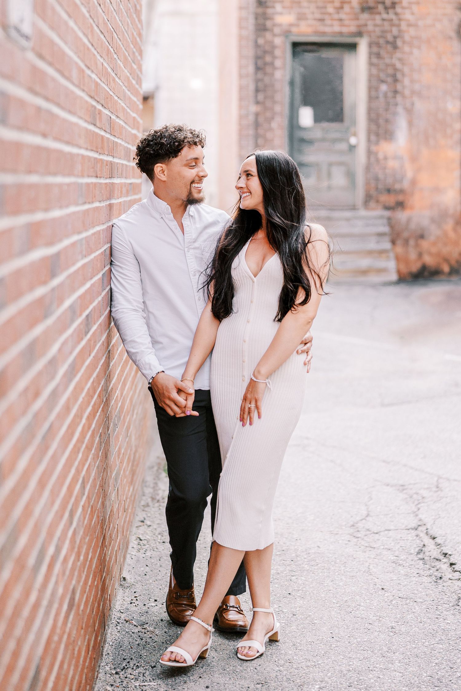 Romantic couple posing against brick wall in elegant attire at urban location.