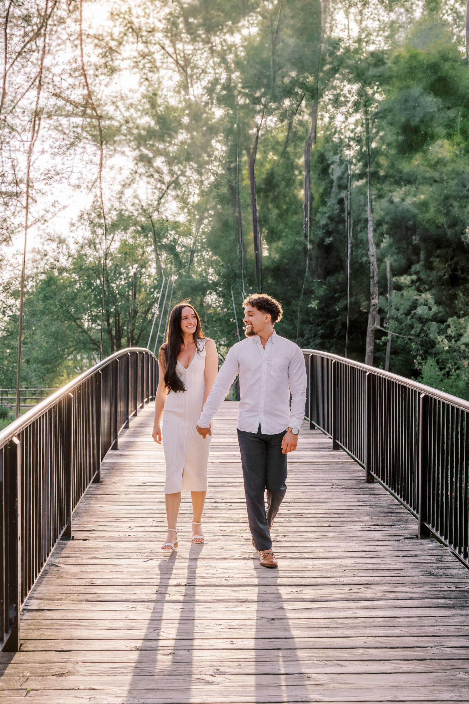 Two figures walking hand in hand on a wooden bridge surrounded by lush green trees in romantic afternoon light.