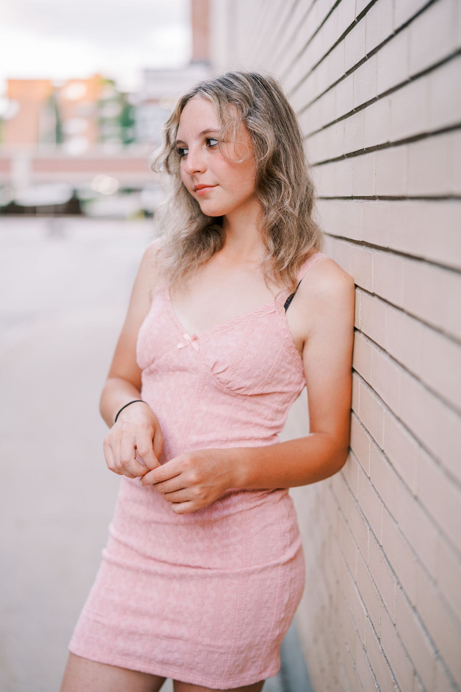 Young woman in pink fitted dress poses against beige wall on sunny day.