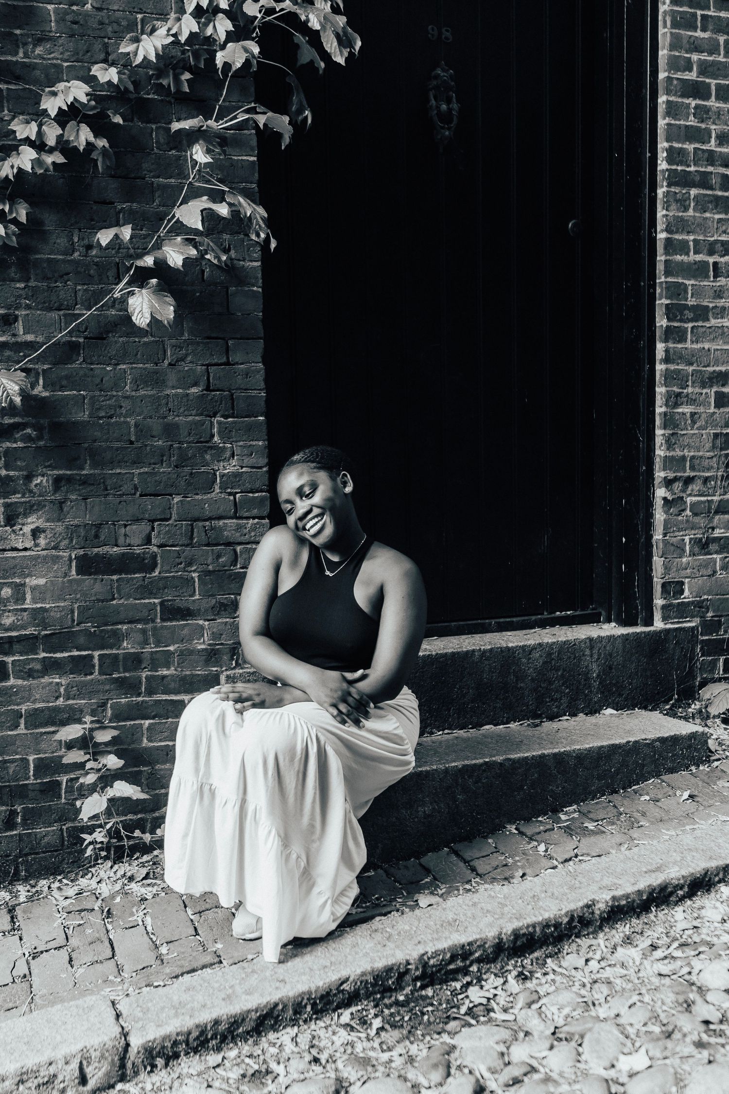 Black and white portrait of someone sitting on brick steps wearing a dark top and light flowing skirt.