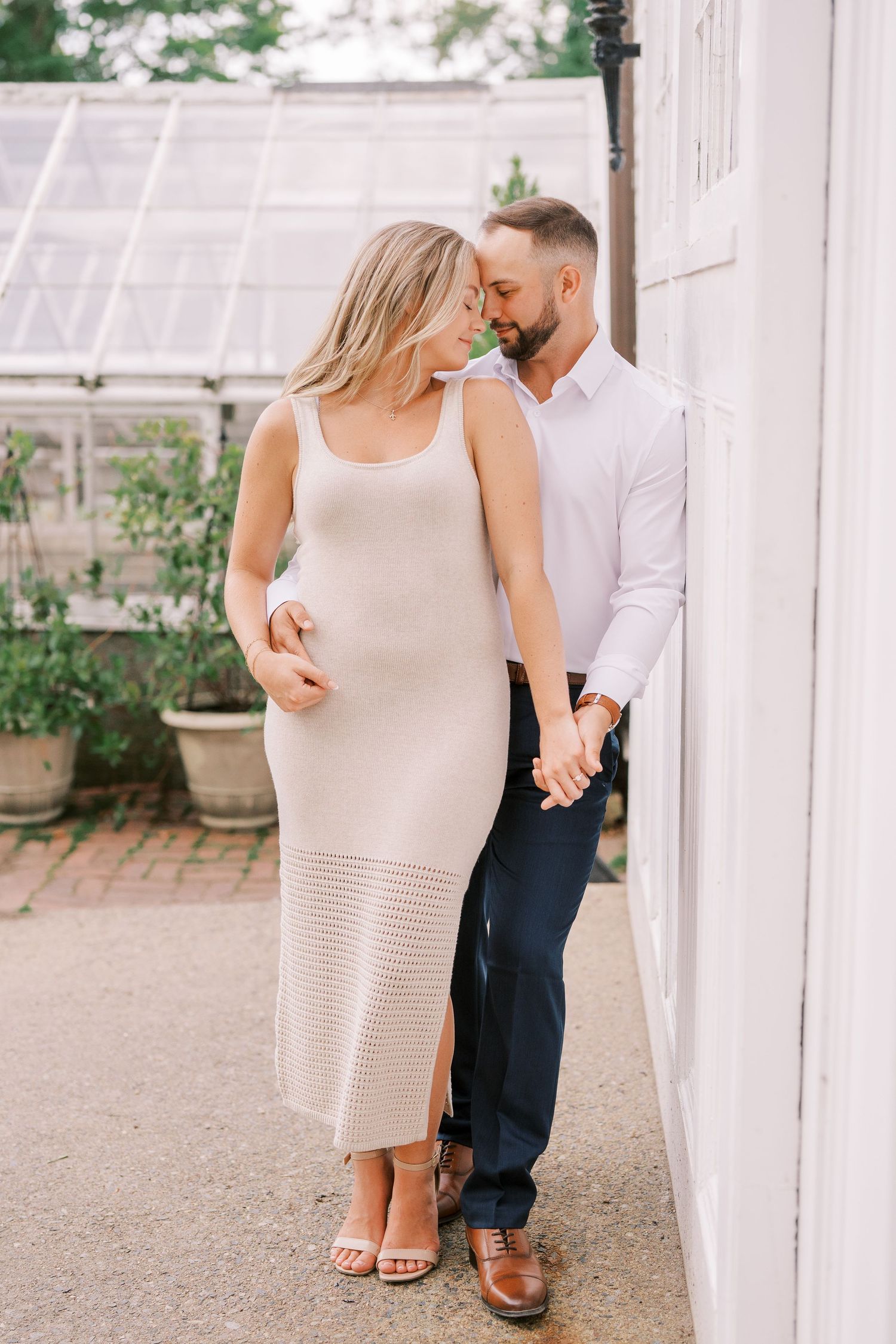 A couple in casual elegant attire share an intimate moment against a white wall near a garden greenhouse.