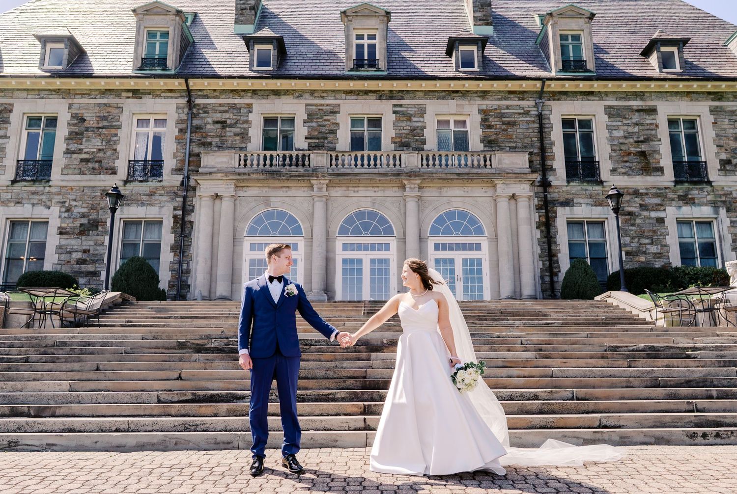 Elegant couple stands on grand stone steps in front of a luxurious French chateau-style mansion.