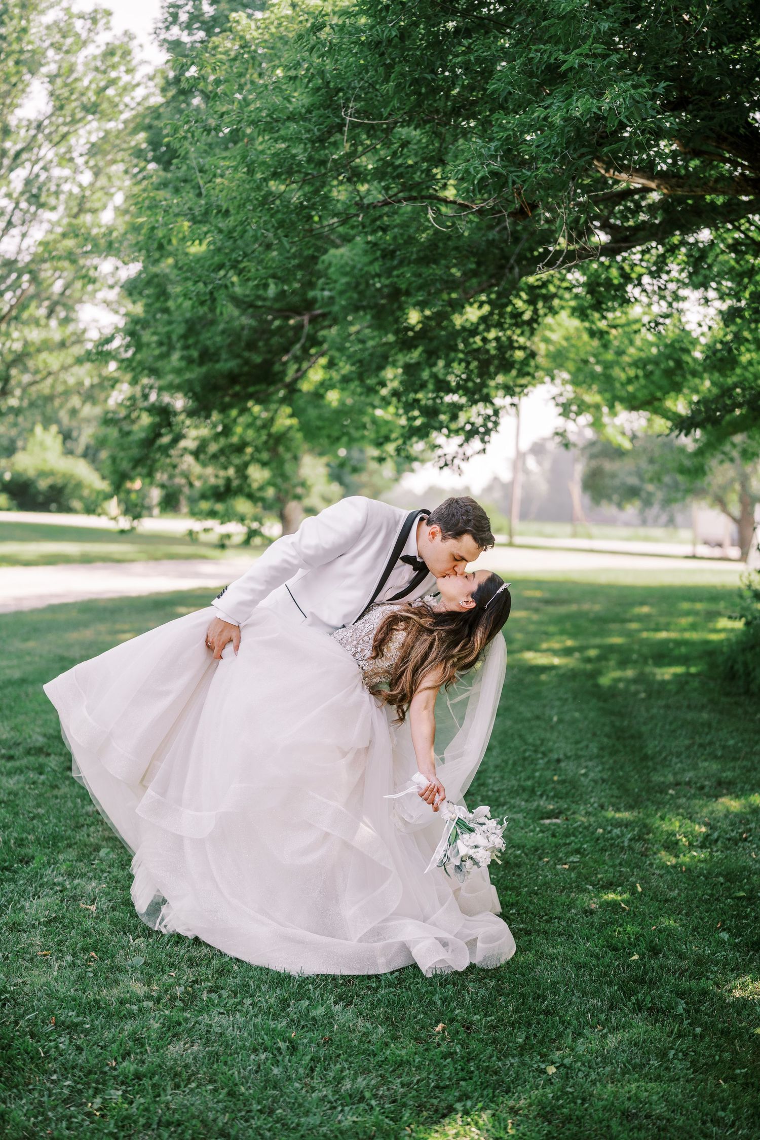 A romantic wedding couple shares a joyful dip pose in a lush green park setting on a sunny day.