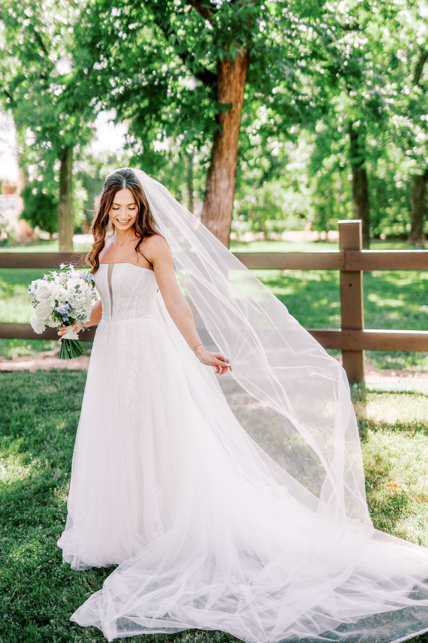 A bride in a flowing white gown with a long veil holds a white bouquet while standing in a sunlit garden setting.