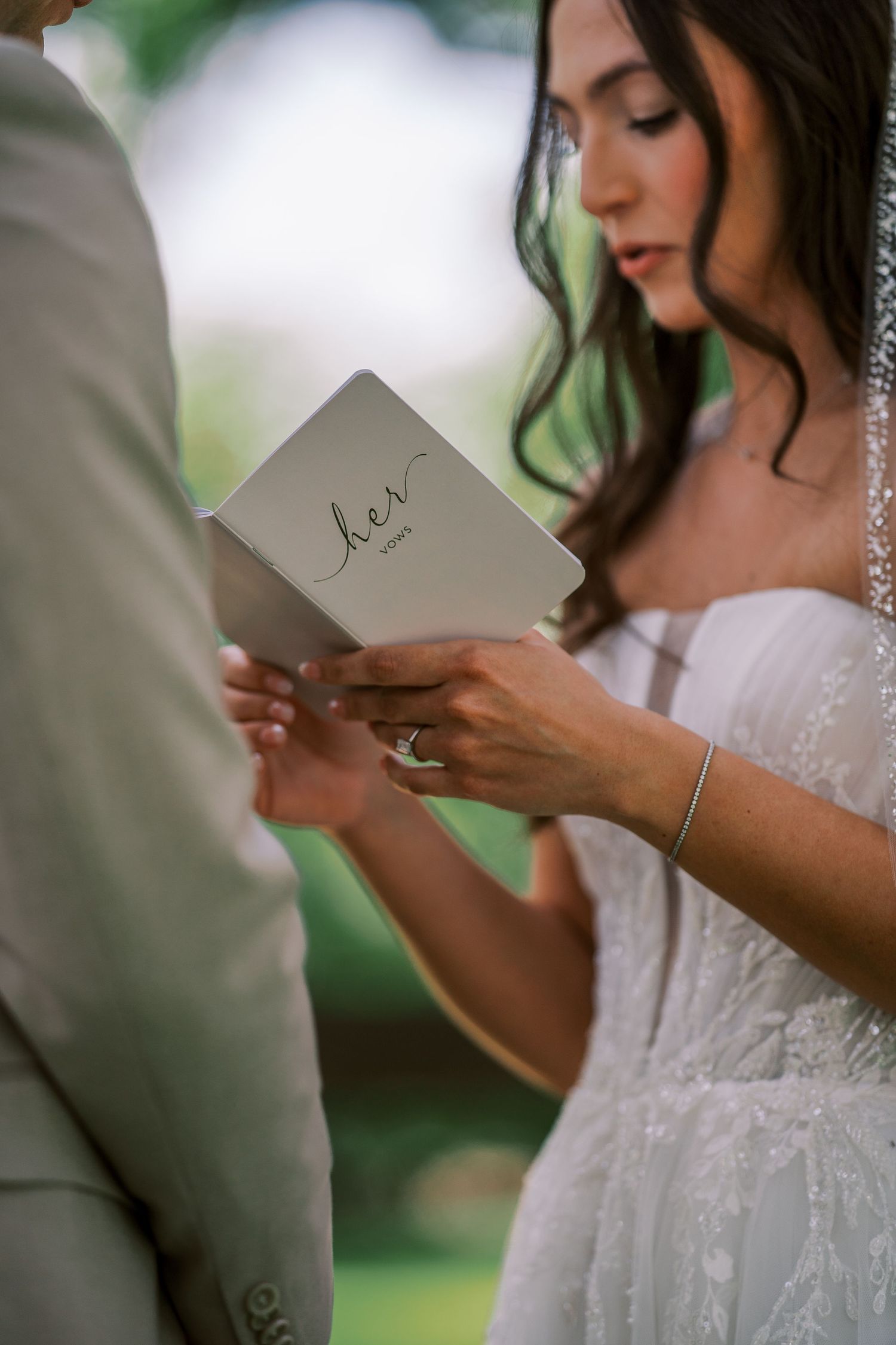 A person in a white lace wedding dress reads handwritten vows during an outdoor wedding ceremony.