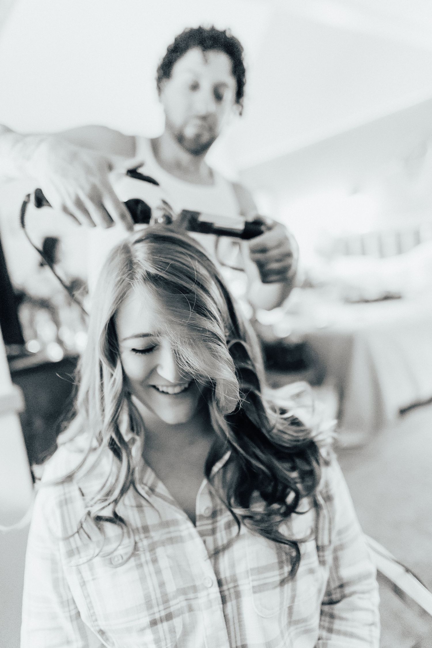 A candid black and white image of a hairstylist styling long wavy hair during wedding preparation.