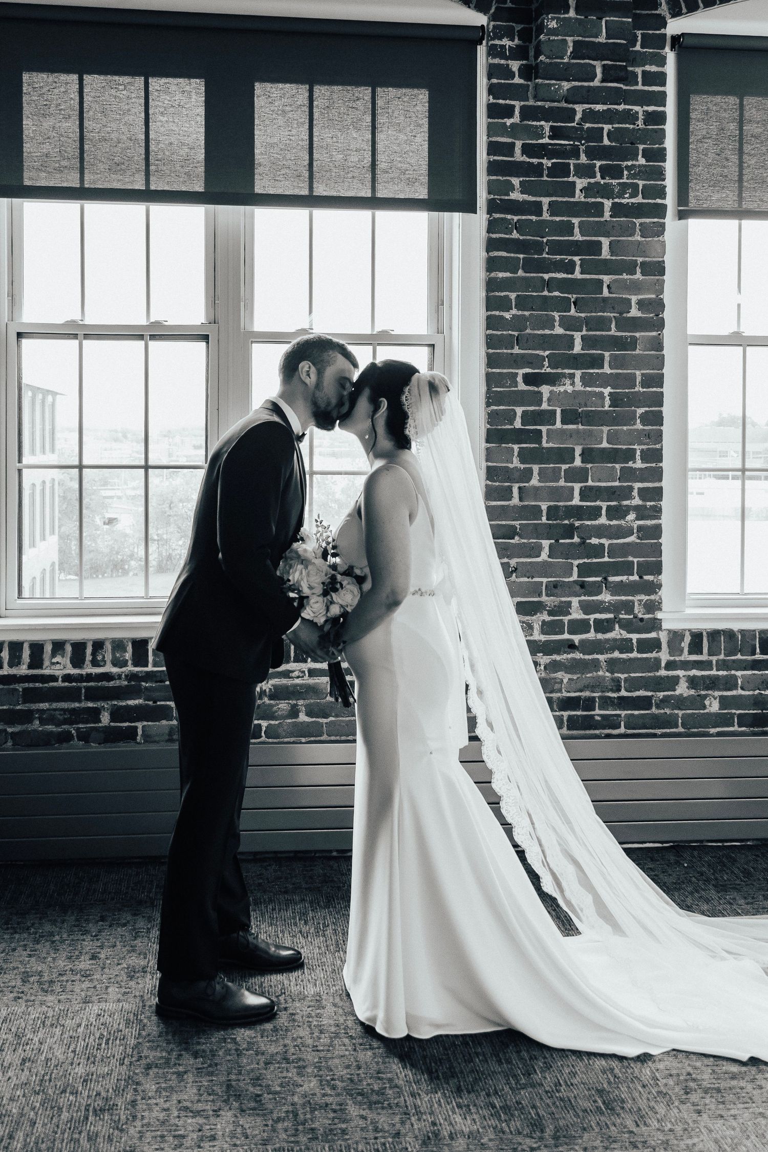 Romantic black and white wedding photo of a couple against exposed brick and large windows.