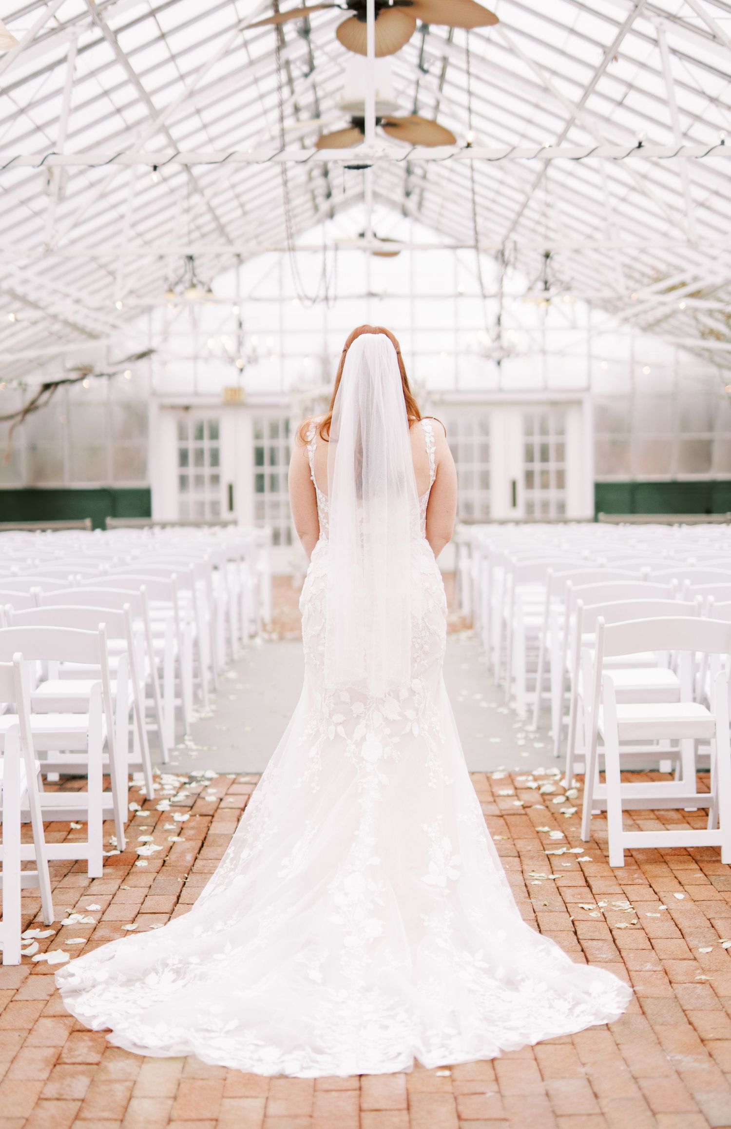 Back view of a bride in a lace wedding gown standing in a greenhouse chapel with white chairs.