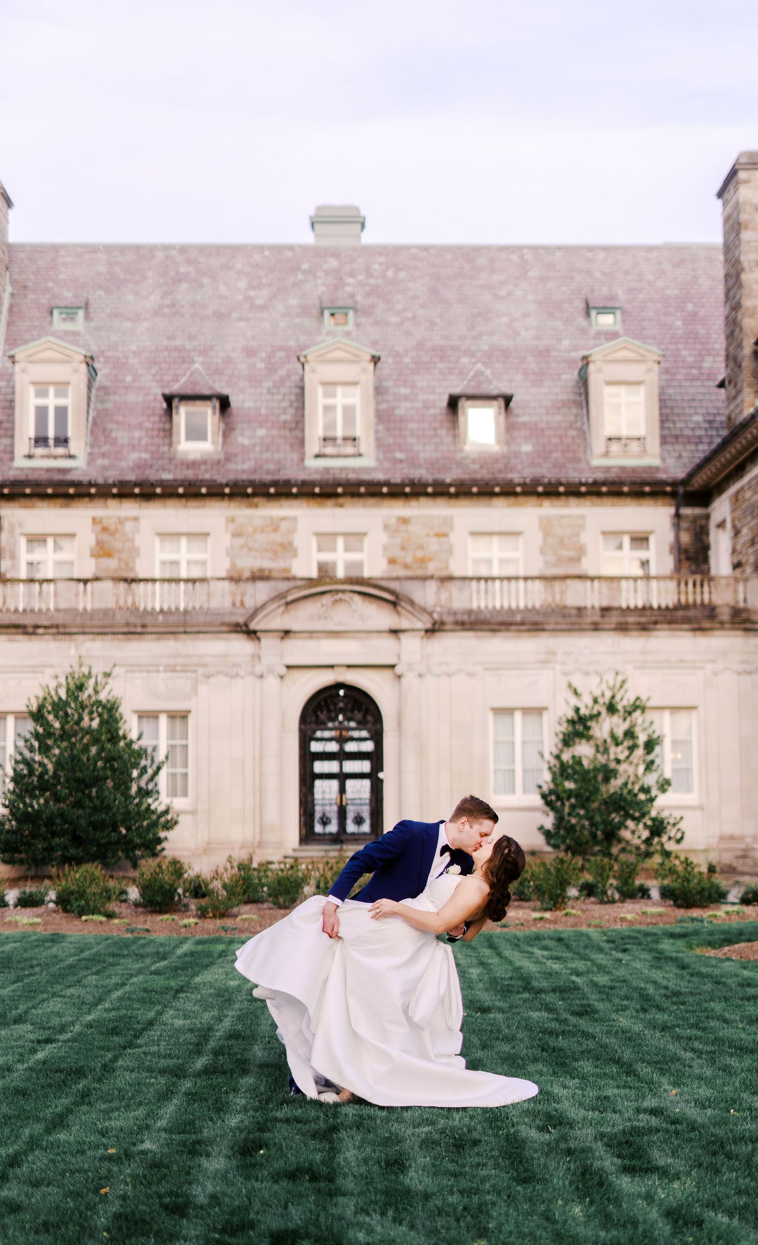 A romantic wedding portrait with the couple embracing on a manicured lawn before a grand historic mansion.