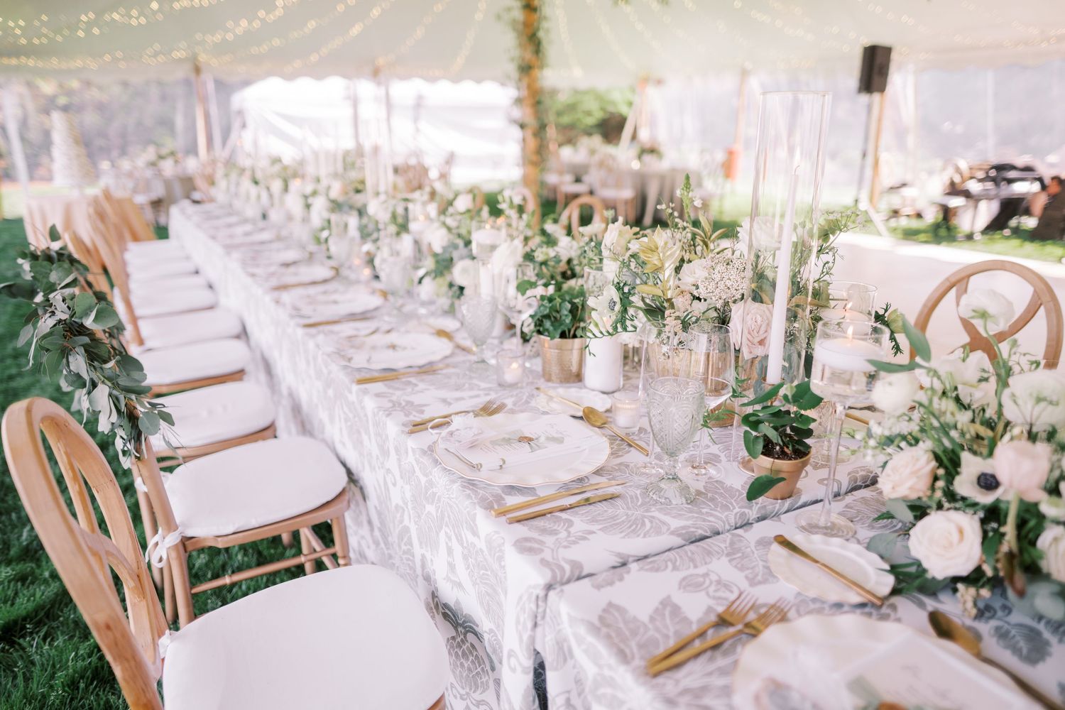 Elegant wedding reception table setting with white linens, gold chargers, and delicate floral centerpieces under a tent.
